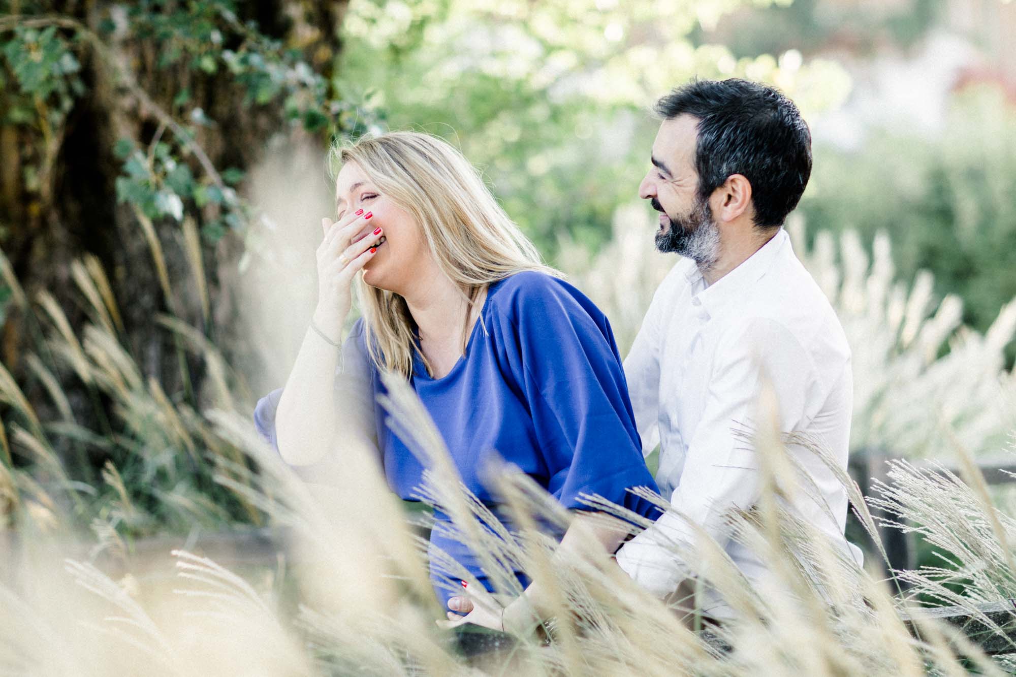 The couple laughing as they sit among the grasses during the maternity session