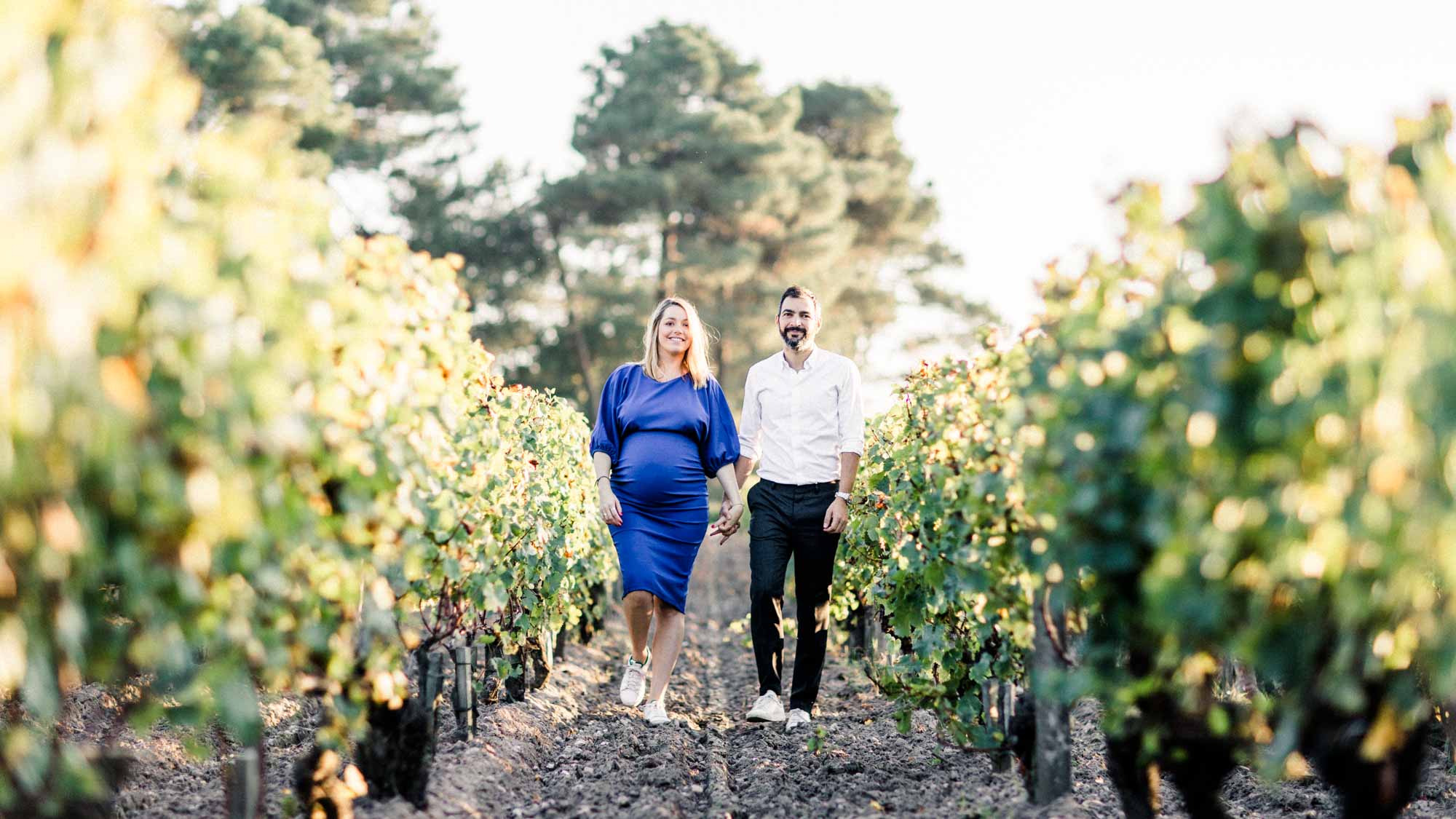 The couple walking down a vine row, the mother-to-be in a blue dress
