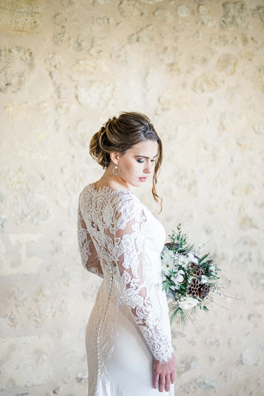 Mariée en robe de dentelle tenant son bouquet devant un mur clair