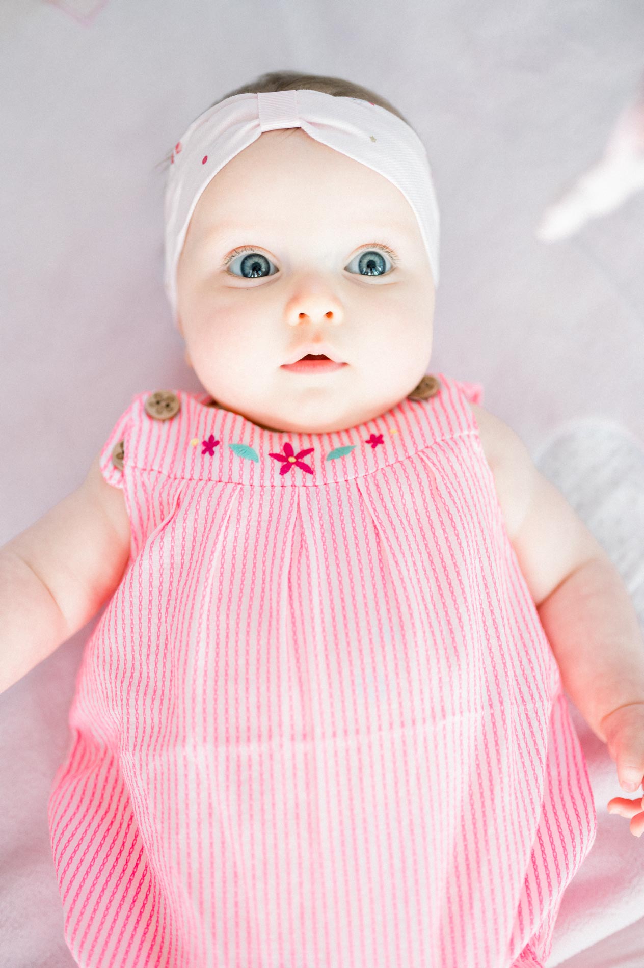 Baby in a pink romper looking surprised, lying on a pale blanket