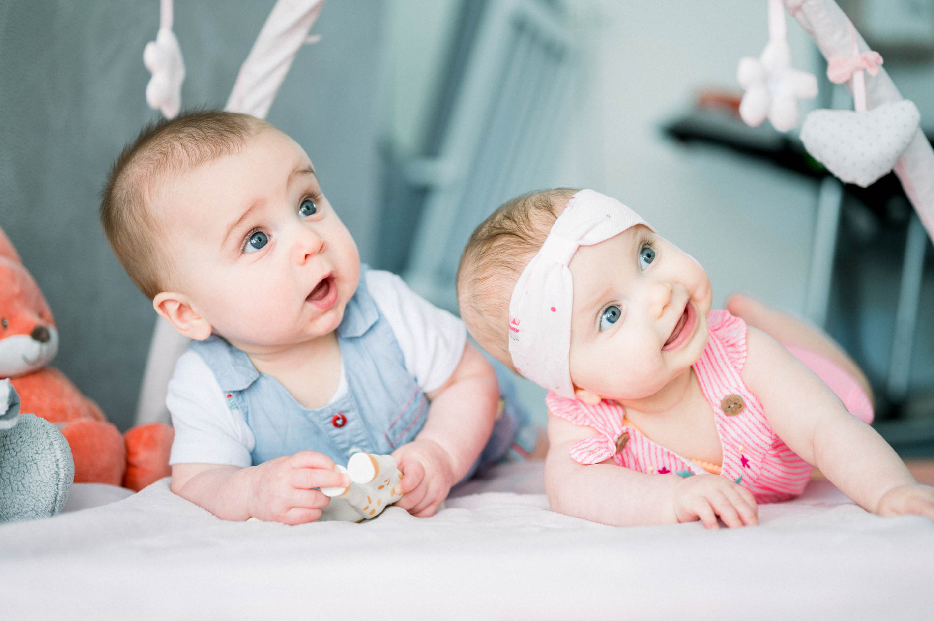 Two babies on their tummies under a play gym, looking curious