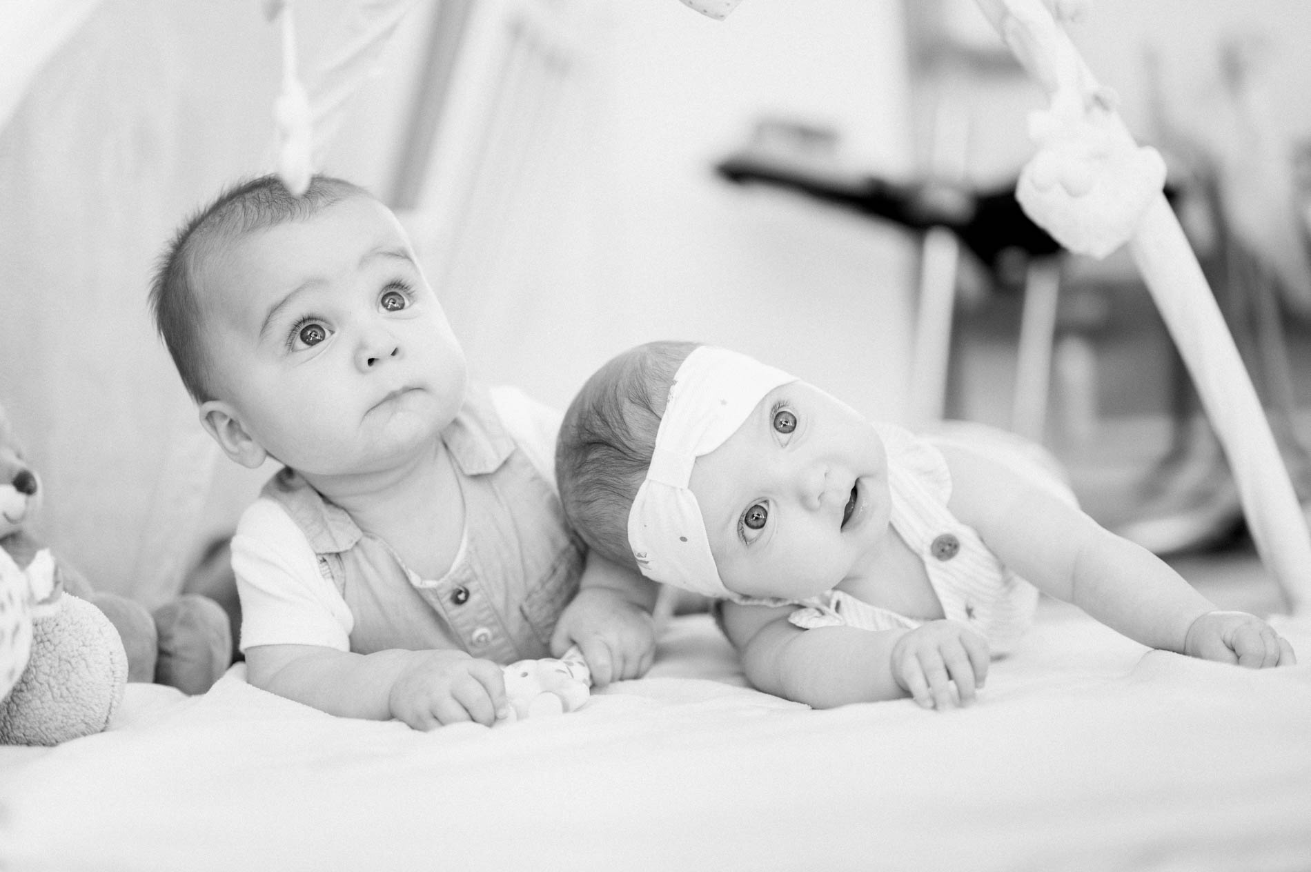 Two babies lying side by side under the play gym, in black and white