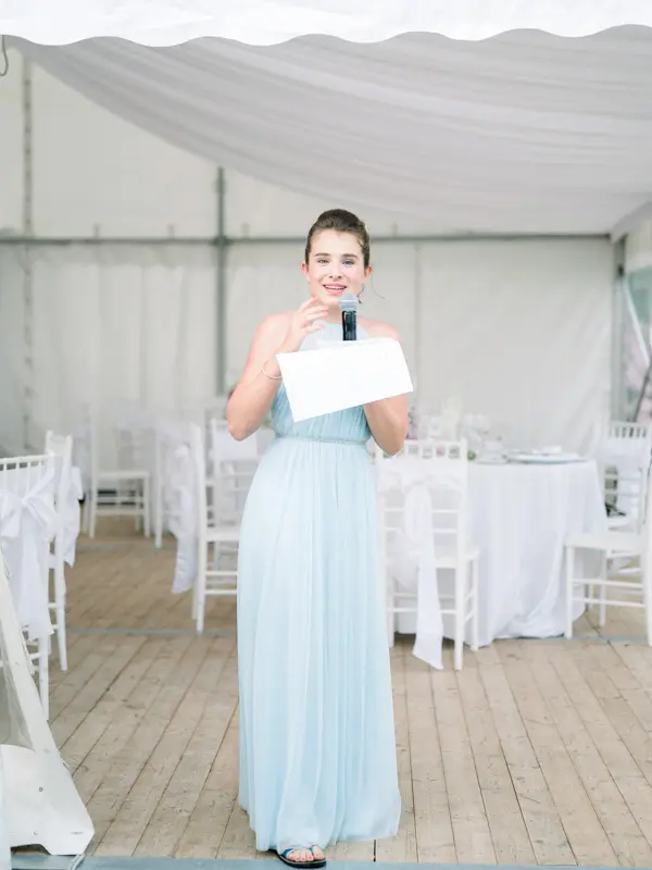 A guest in a pale blue dress gives a speech in the marquee during the cocktail hour