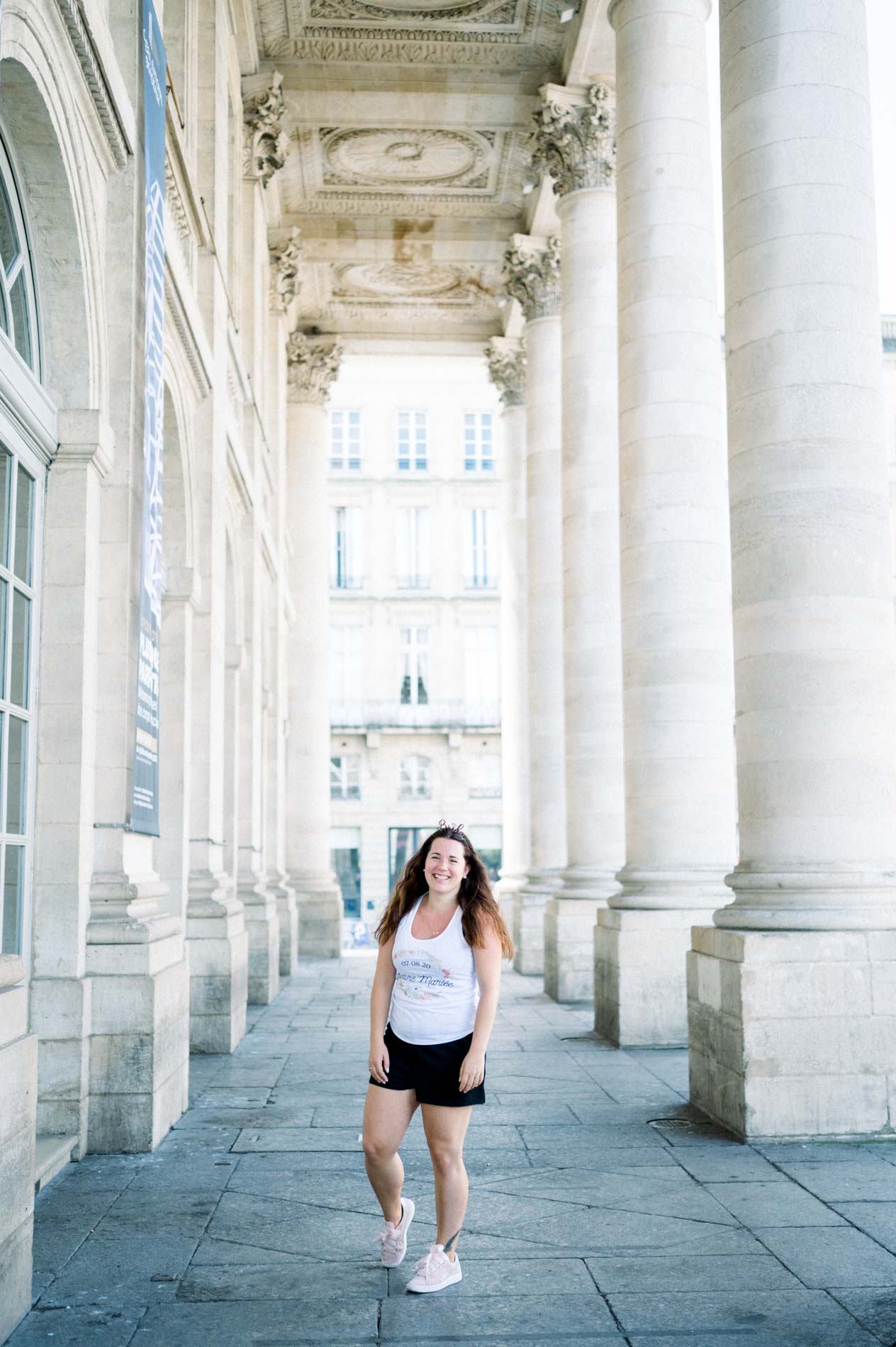 The bride-to-be walking alone beneath a tall stone colonnade in Bordeaux