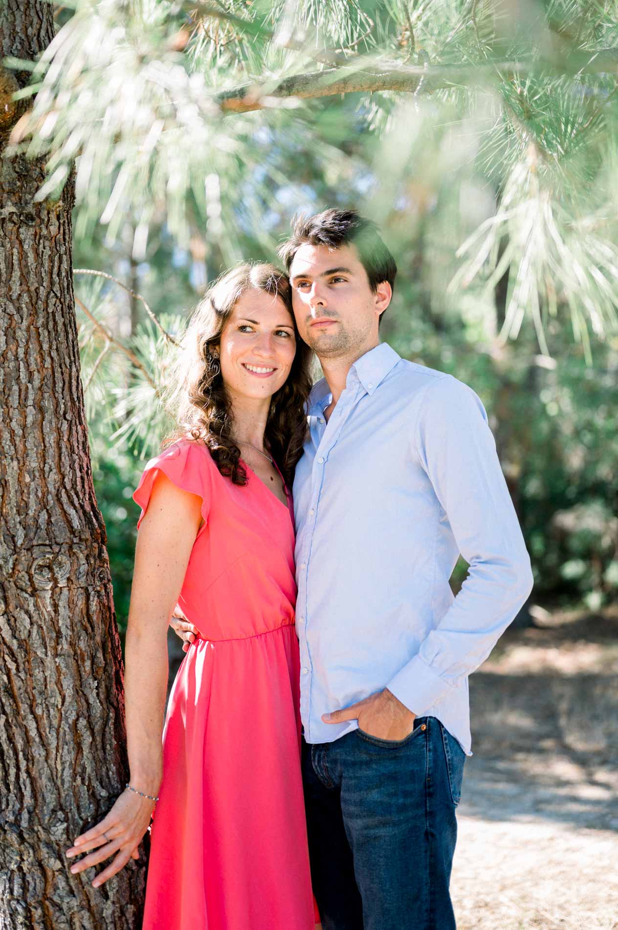 The engaged couple leaning against a pine trunk, she in coral, he in a blue shirt