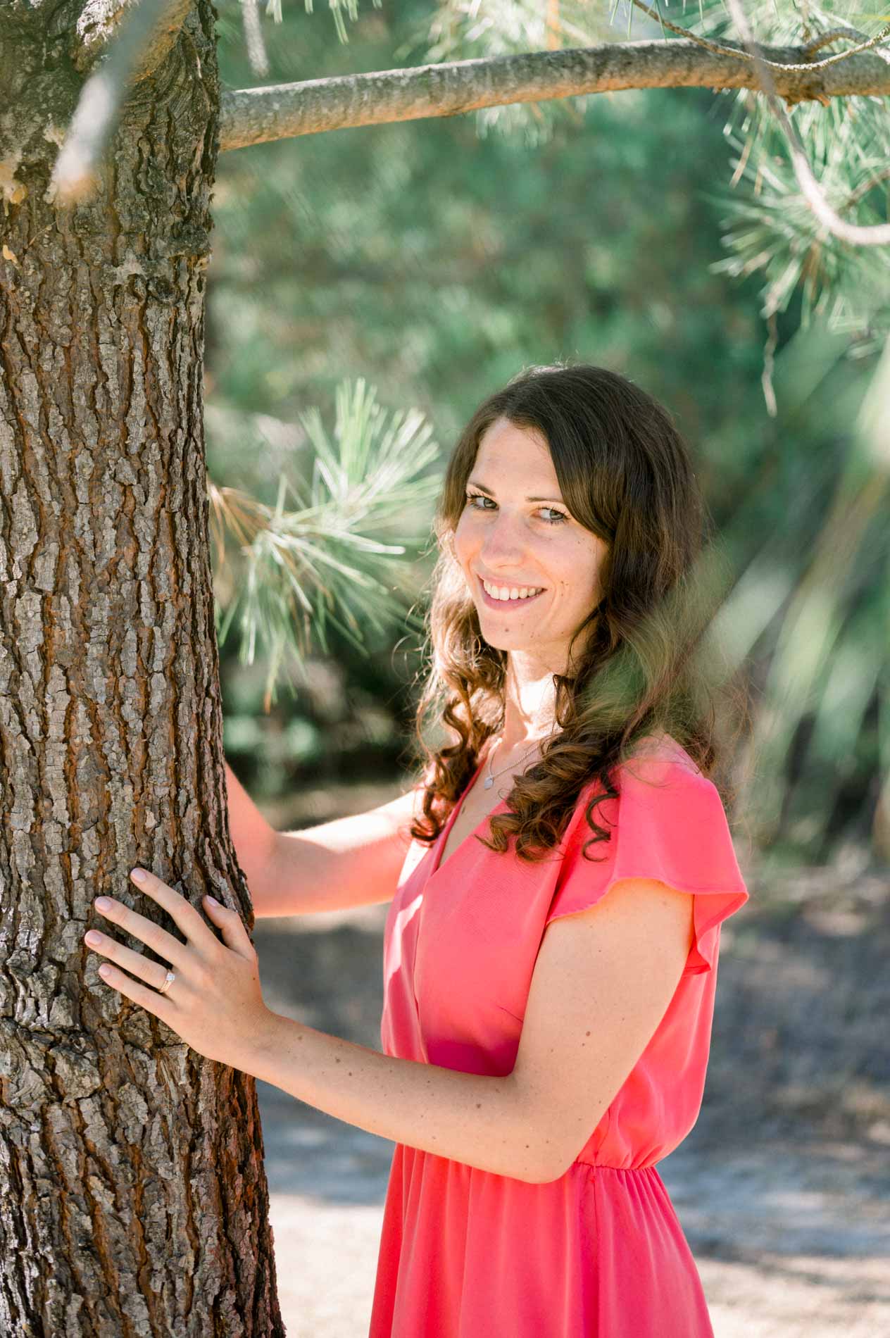 The bride-to-be in a coral dress smiling with her hand on a pine tree
