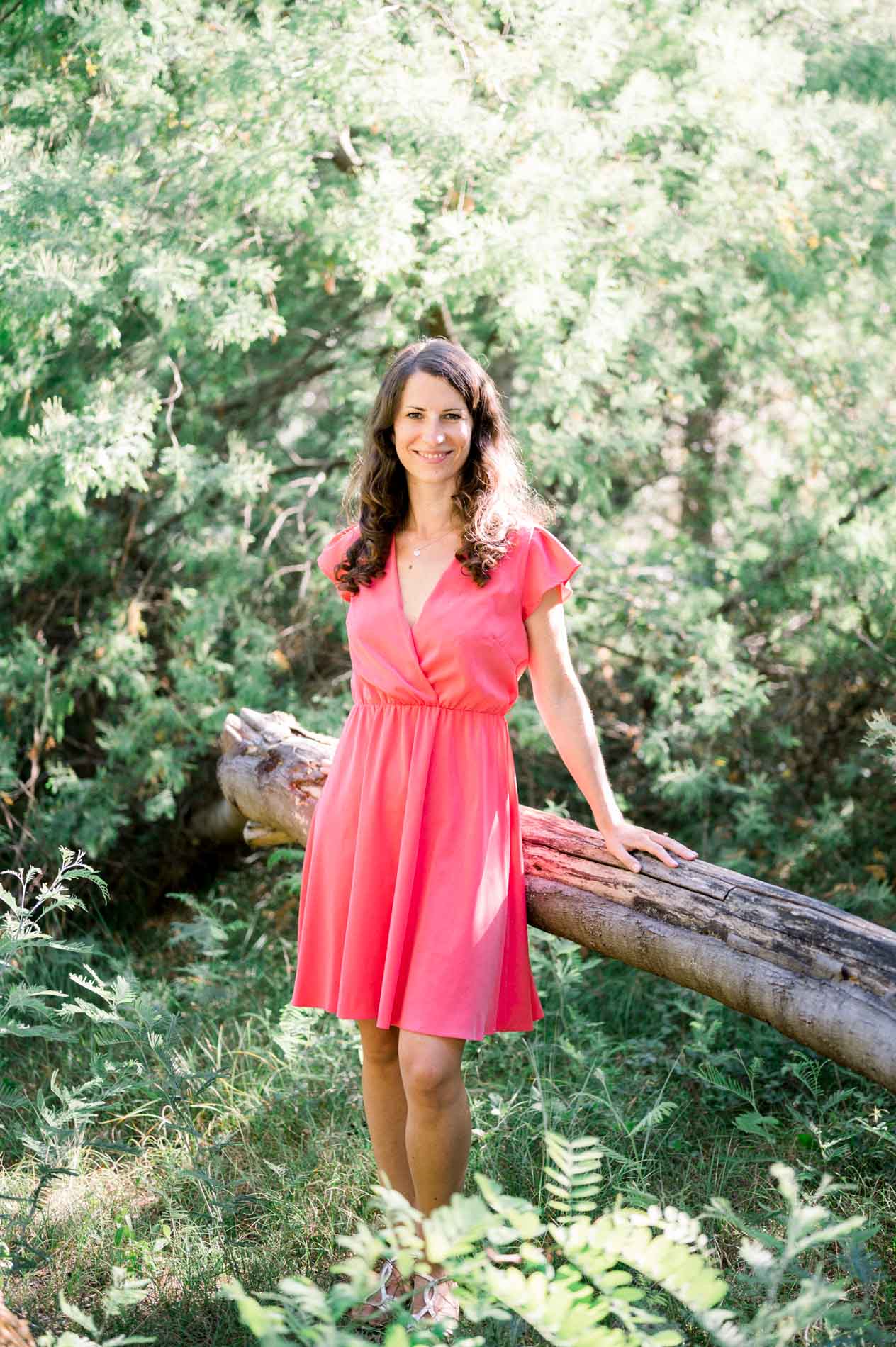 The bride-to-be in a coral dress standing by a fallen trunk in the pine forest