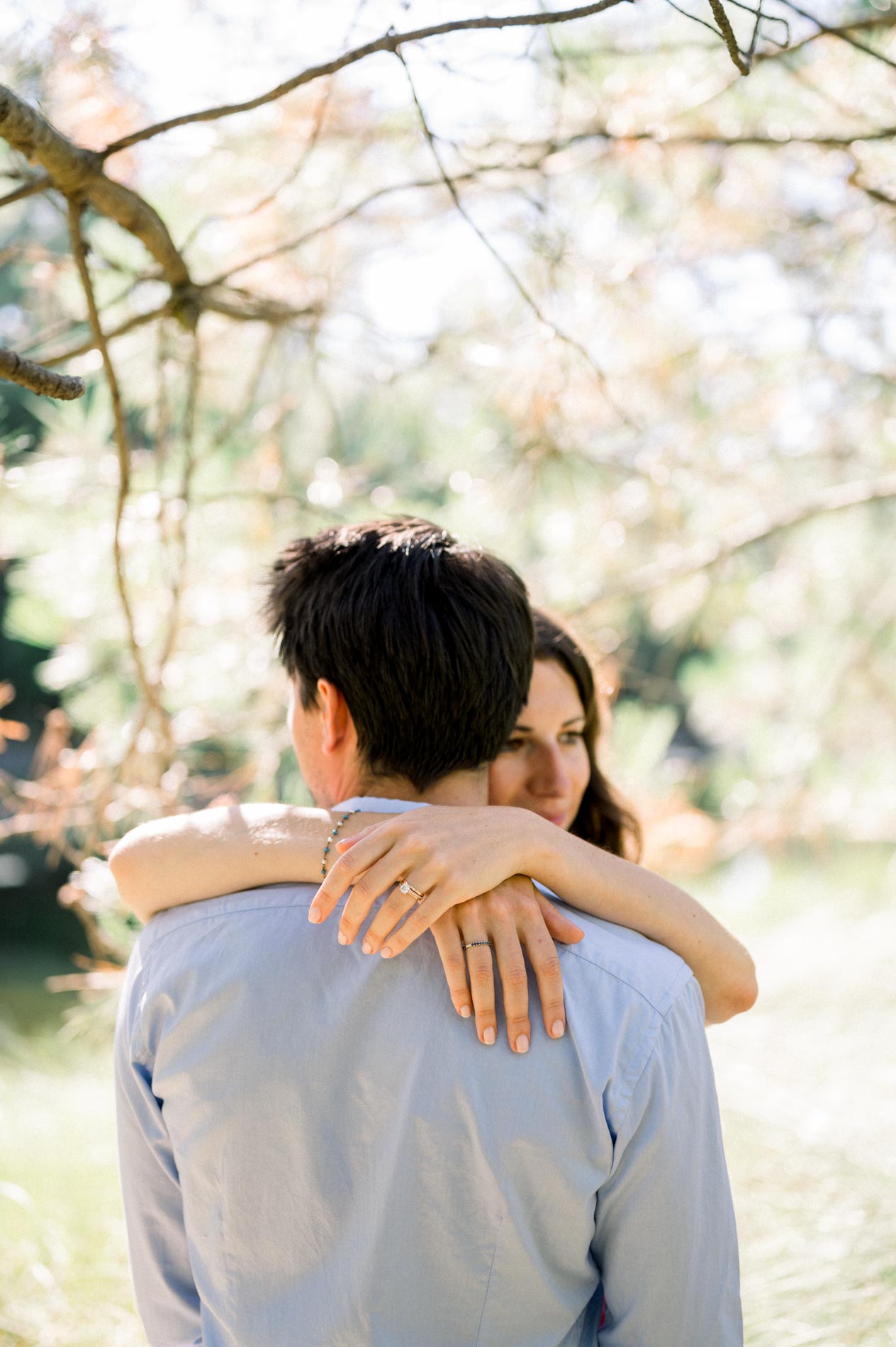 The bride-to-be wraps her arms around her fiancé from behind under the branches