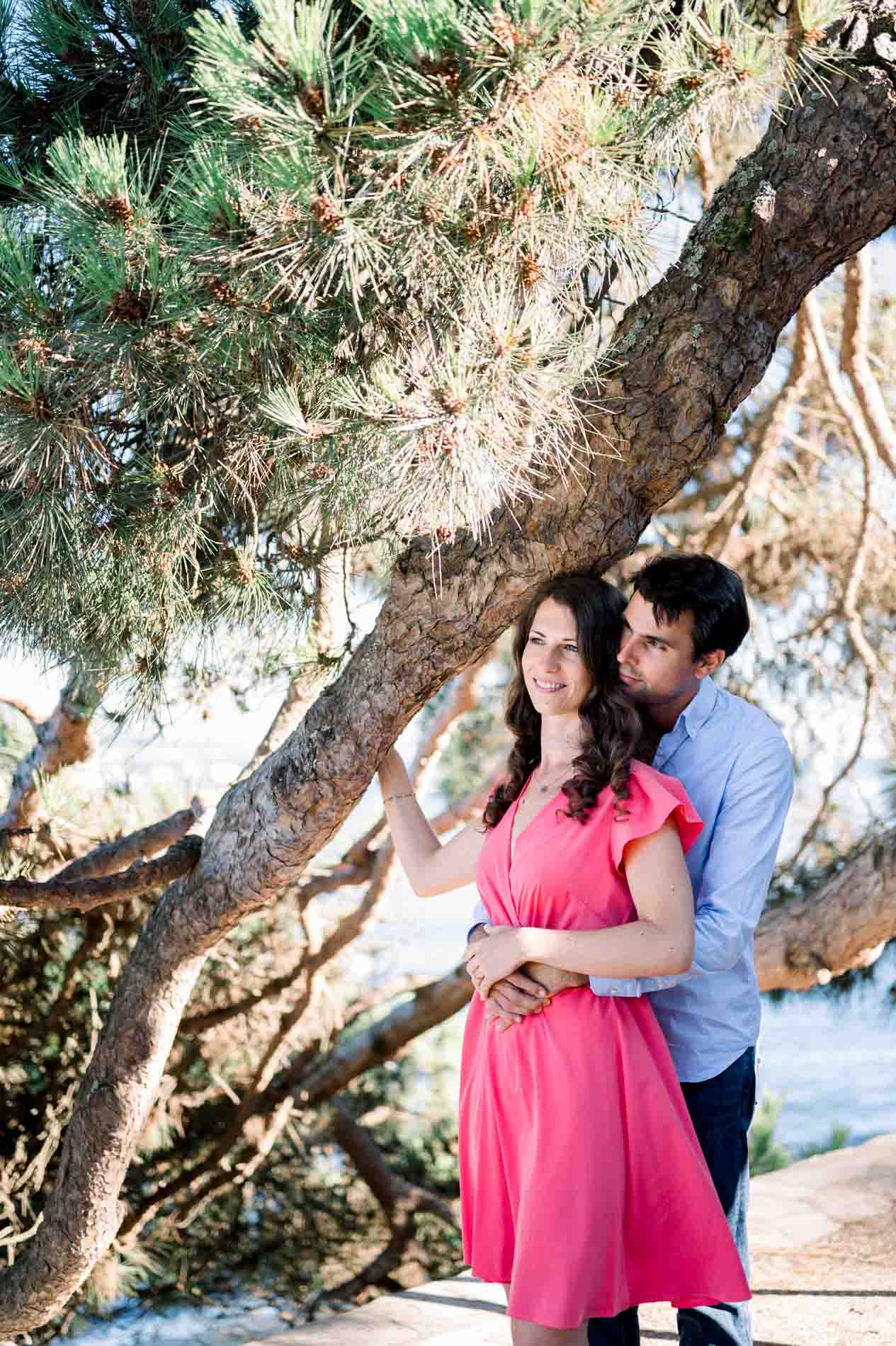 The fiancé holds the bride-to-be under a leaning pine by the bay