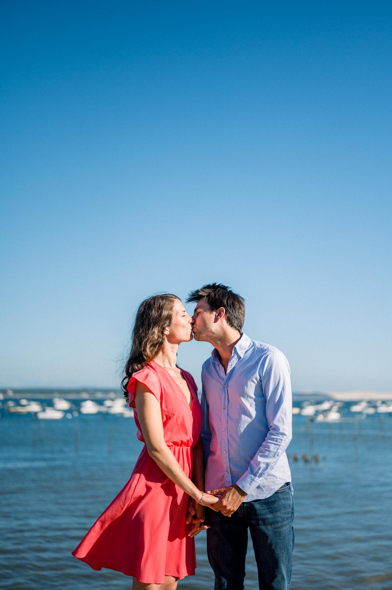 The couple kiss on the shore of Arcachon Bay with moored boats behind them