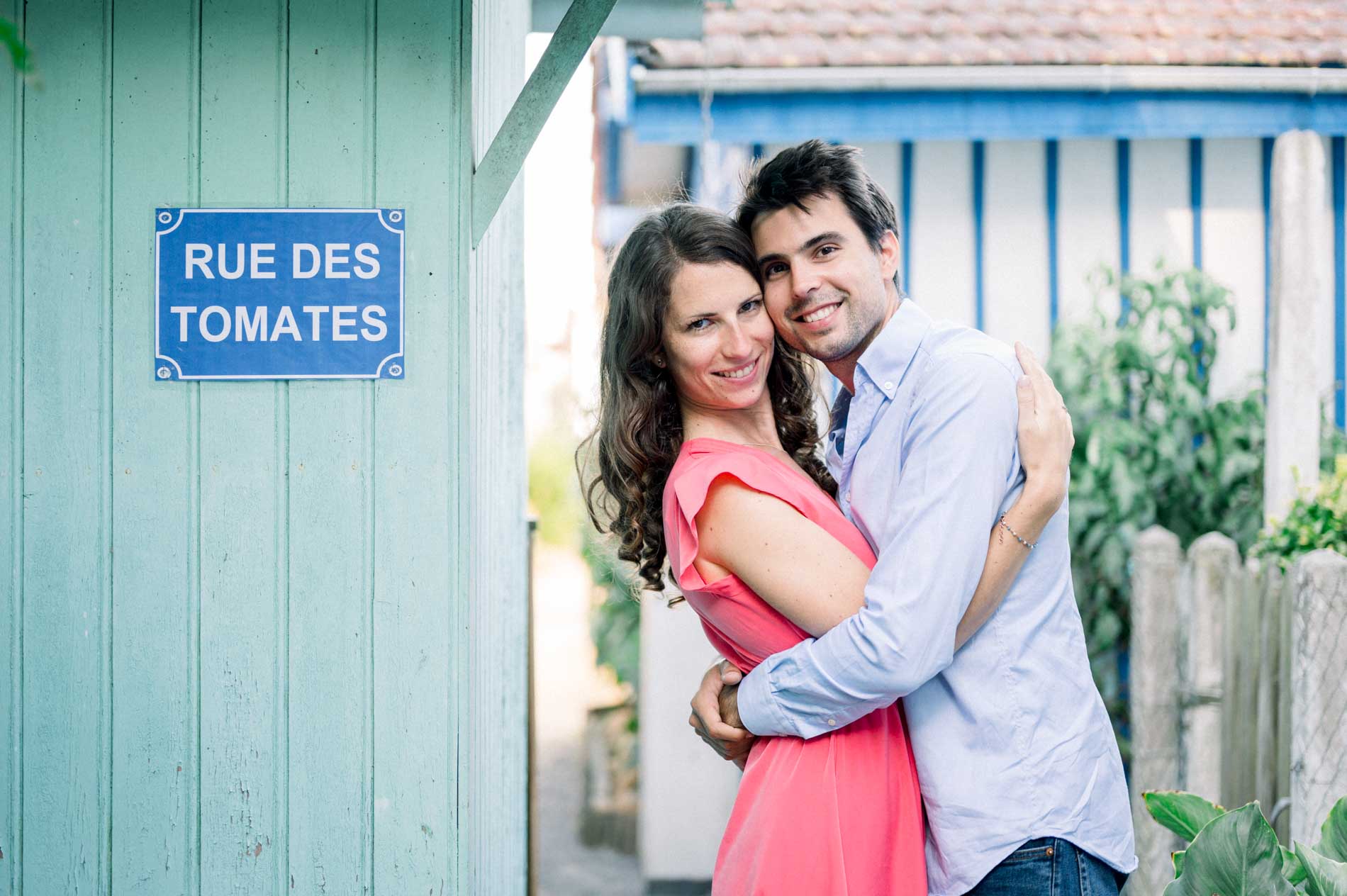 The engaged couple embracing by the “Rue des Tomates” sign on a blue hut