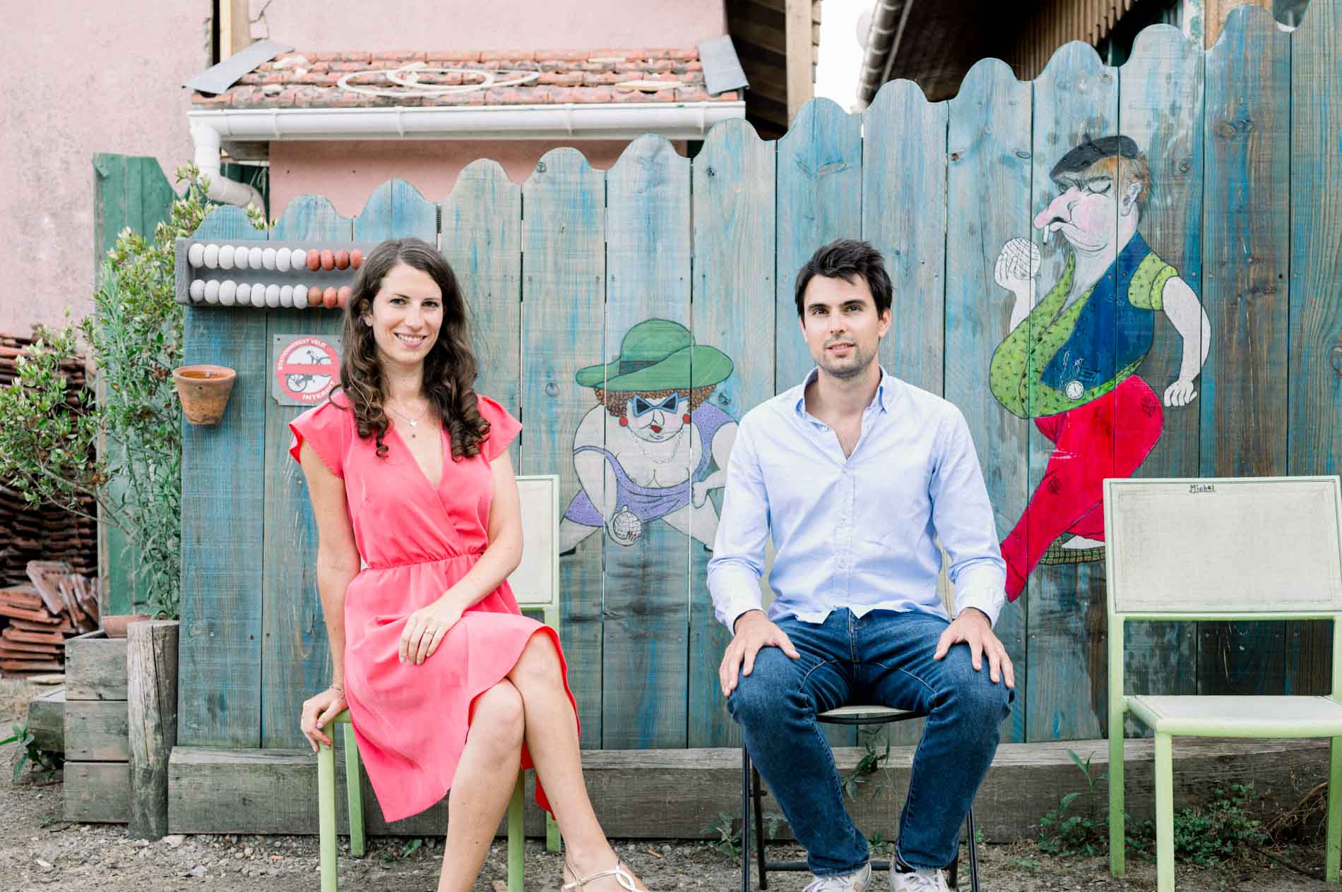 The couple seated on chairs in front of a fence painted with colourful characters