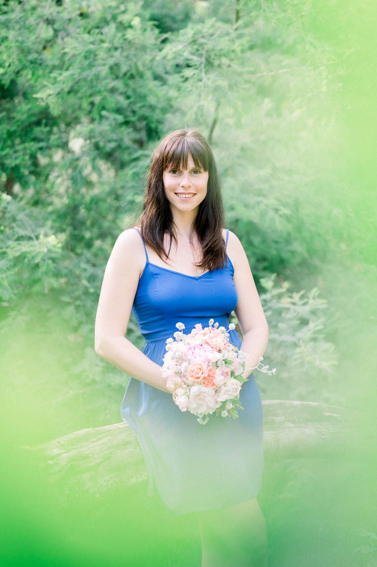 The bride-to-be in a blue dress holding her bouquet, framed by foliage