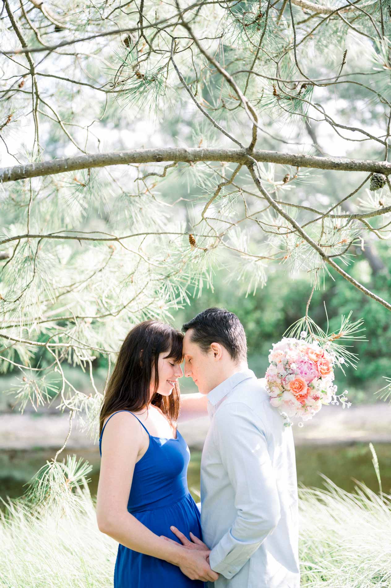 The engaged couple face to face beneath pine branches by the water