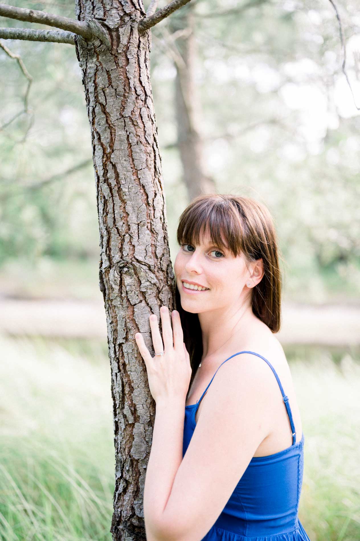 The bride-to-be leaning against a pine trunk in a blue dress, softly smiling