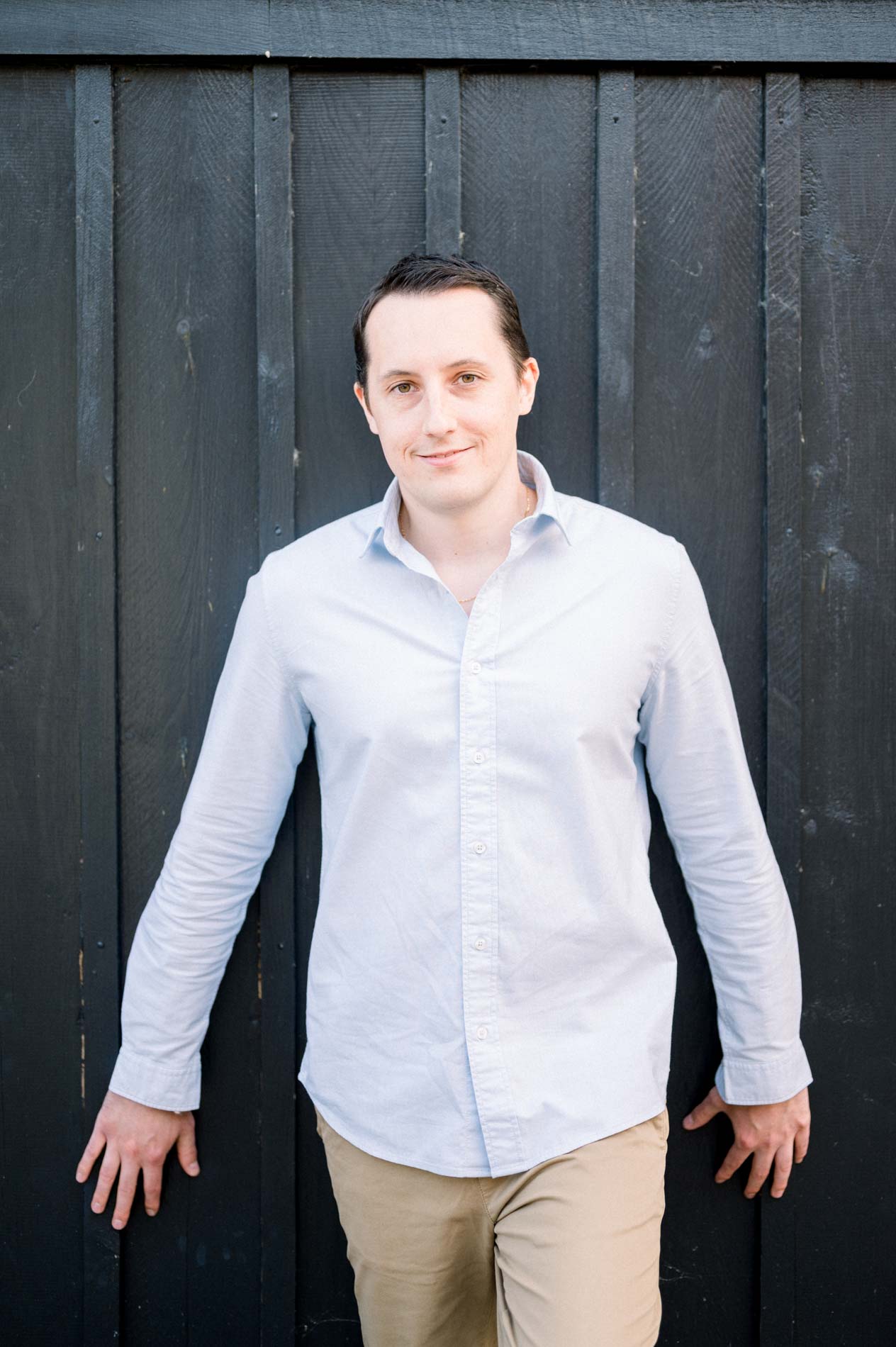 The fiancé in a light shirt leaning against a black wooden cabin