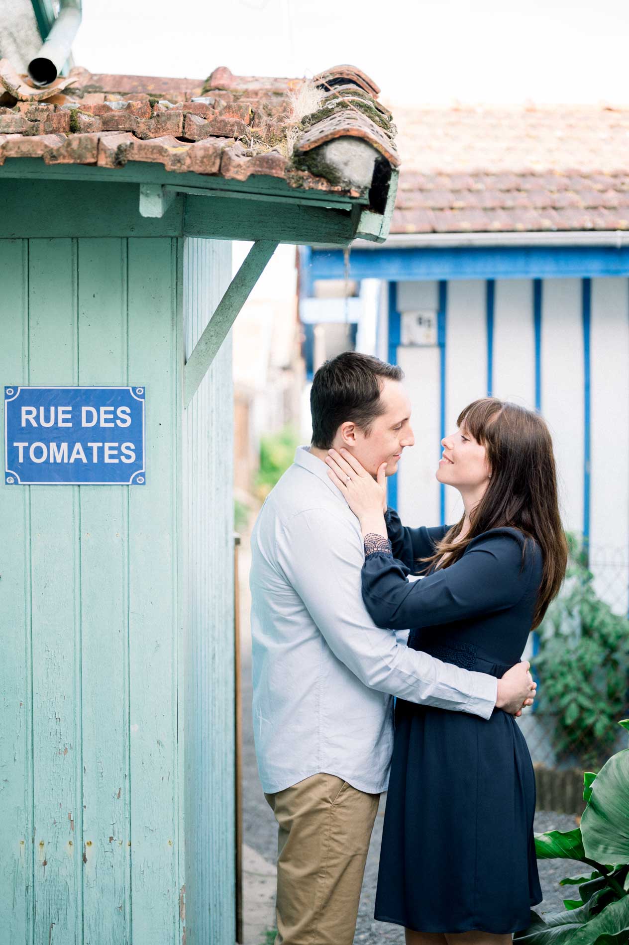 The engaged couple embracing by the “Rue des Tomates” sign and a green cabin