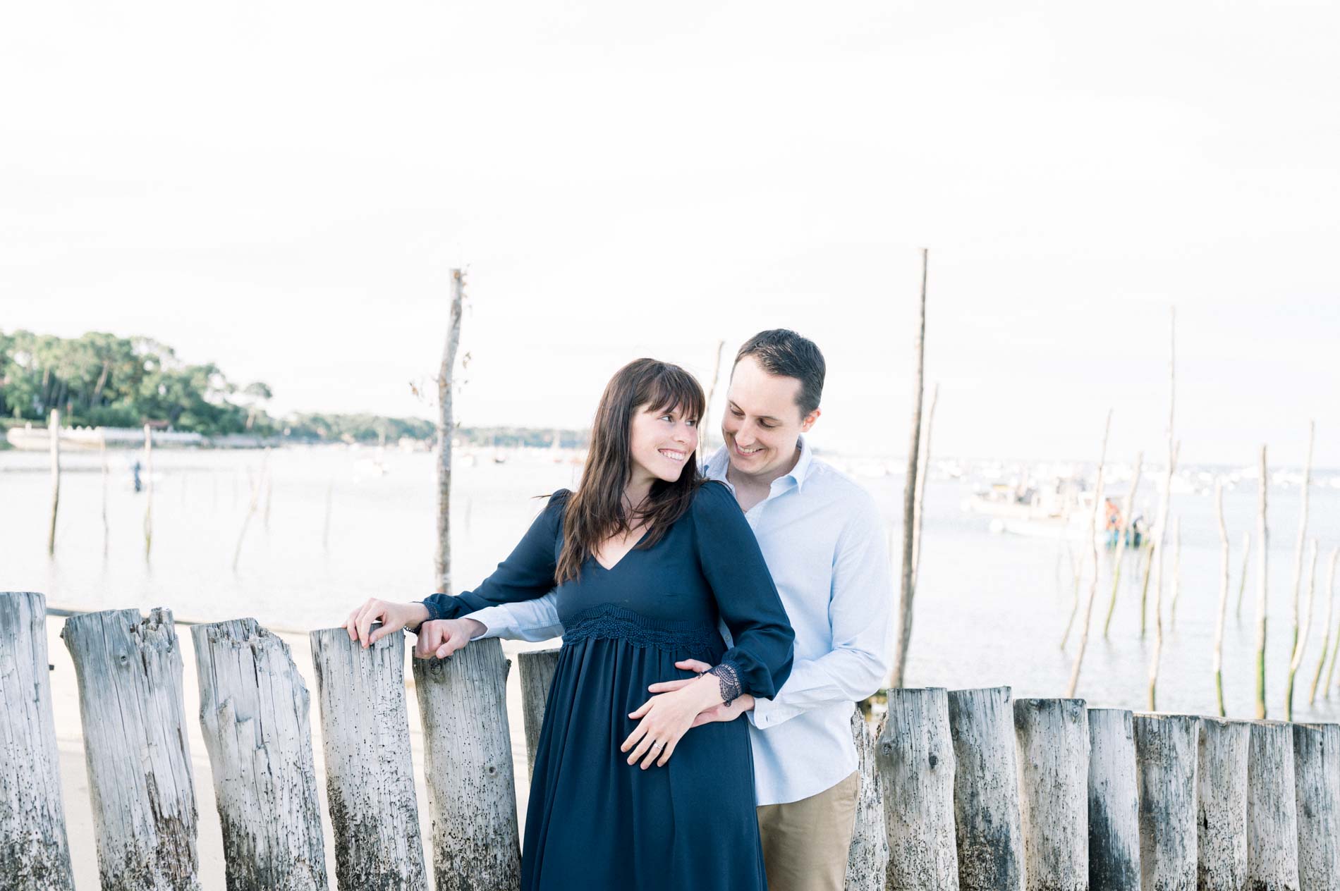 The engaged couple leaning on a wooden stake fence facing Arcachon Bay