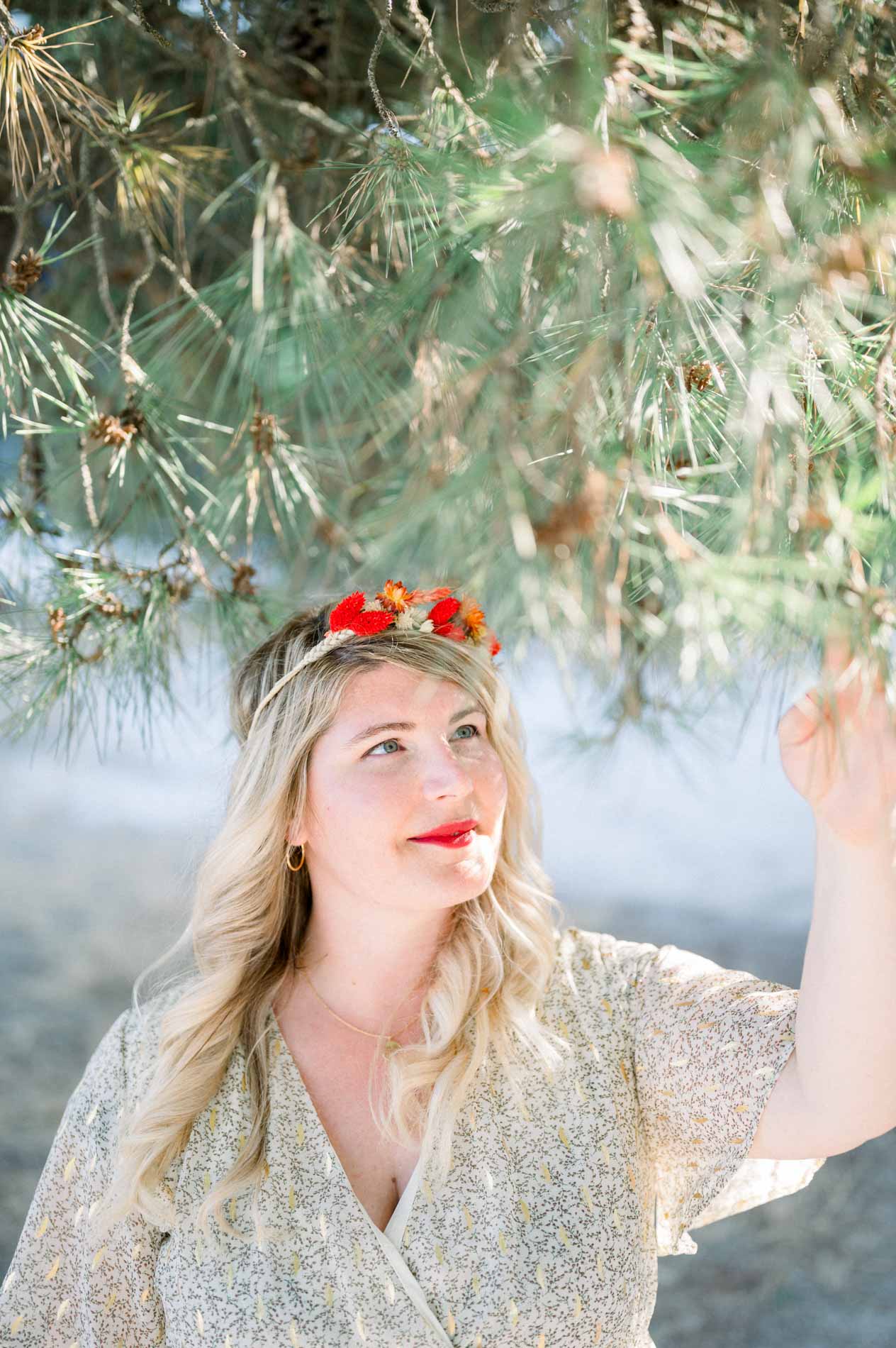 Blonde woman wearing a red flower crown beneath pine branches