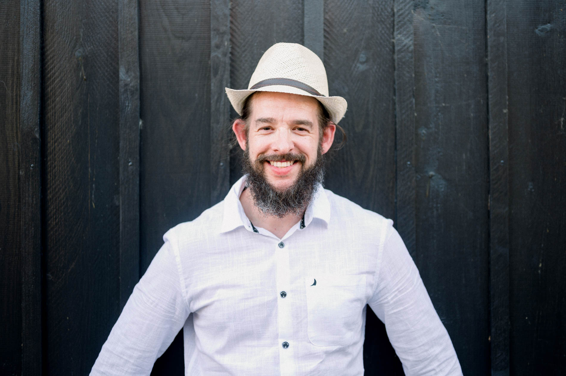 Bearded man in a straw hat smiling in front of a black wooden hut