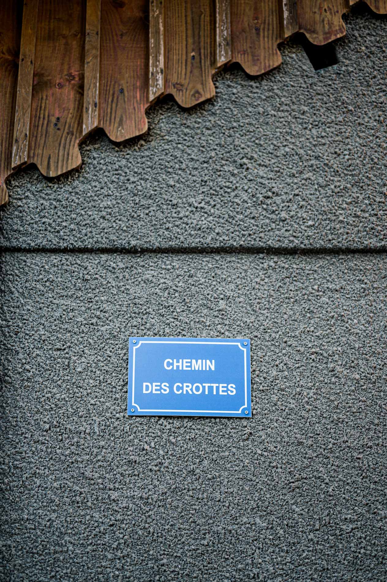 Blue Chemin des Crottes street sign on a grey wall beneath a wooden roof