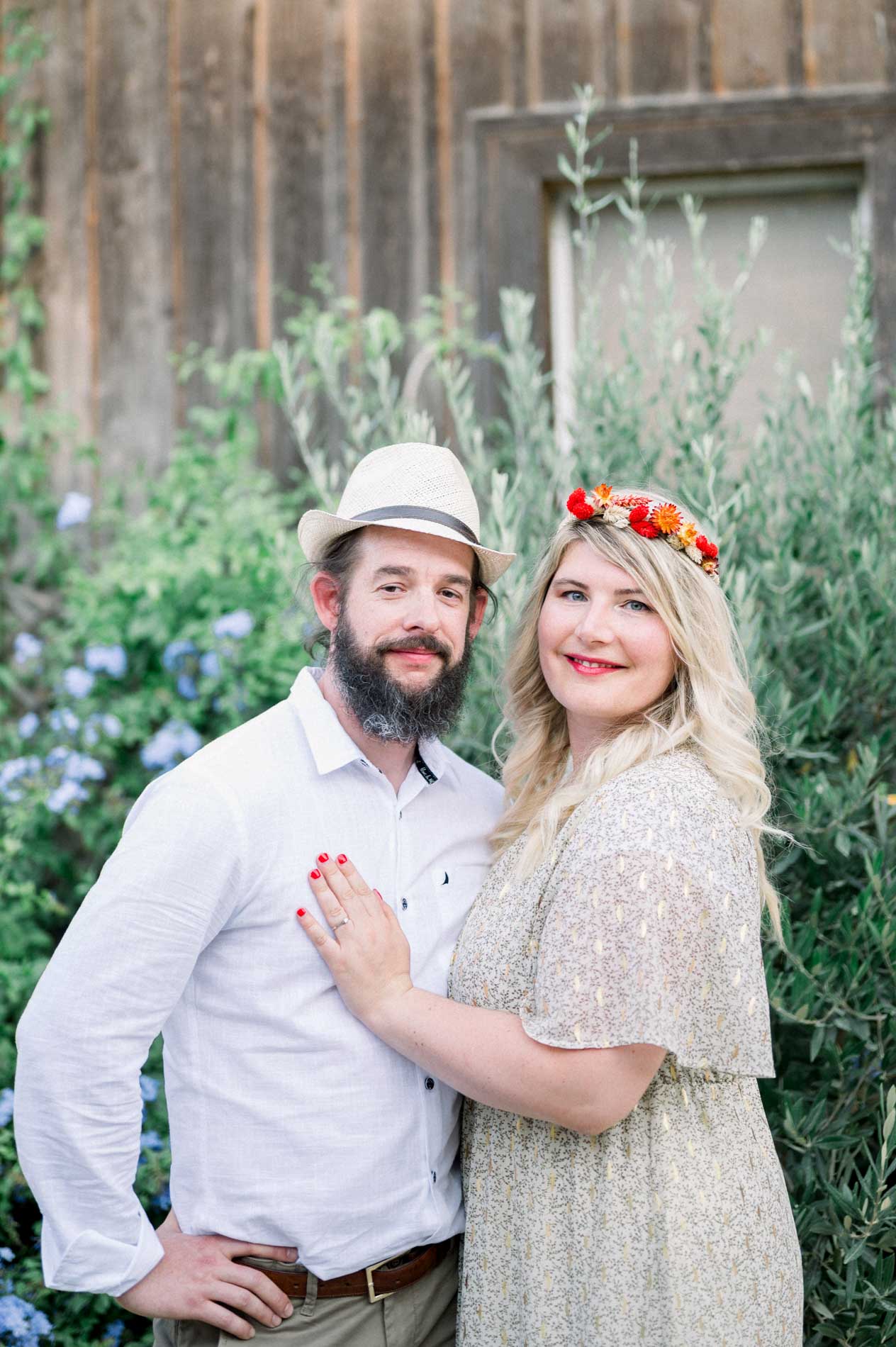 The couple posing by a wooden cabin surrounded by blue flowers