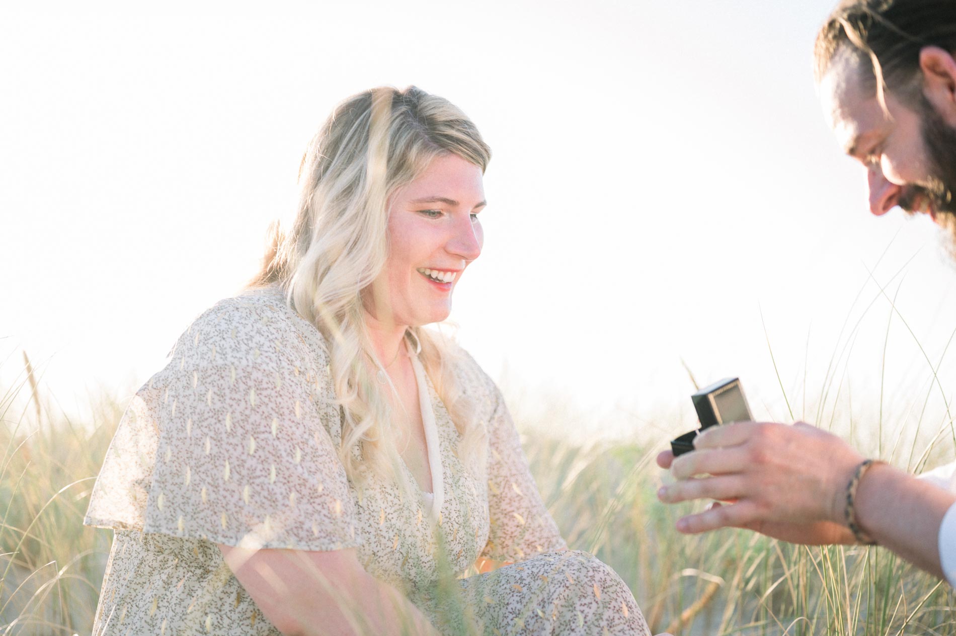 Derek opening the ring box in front of a moved Julie on the beach