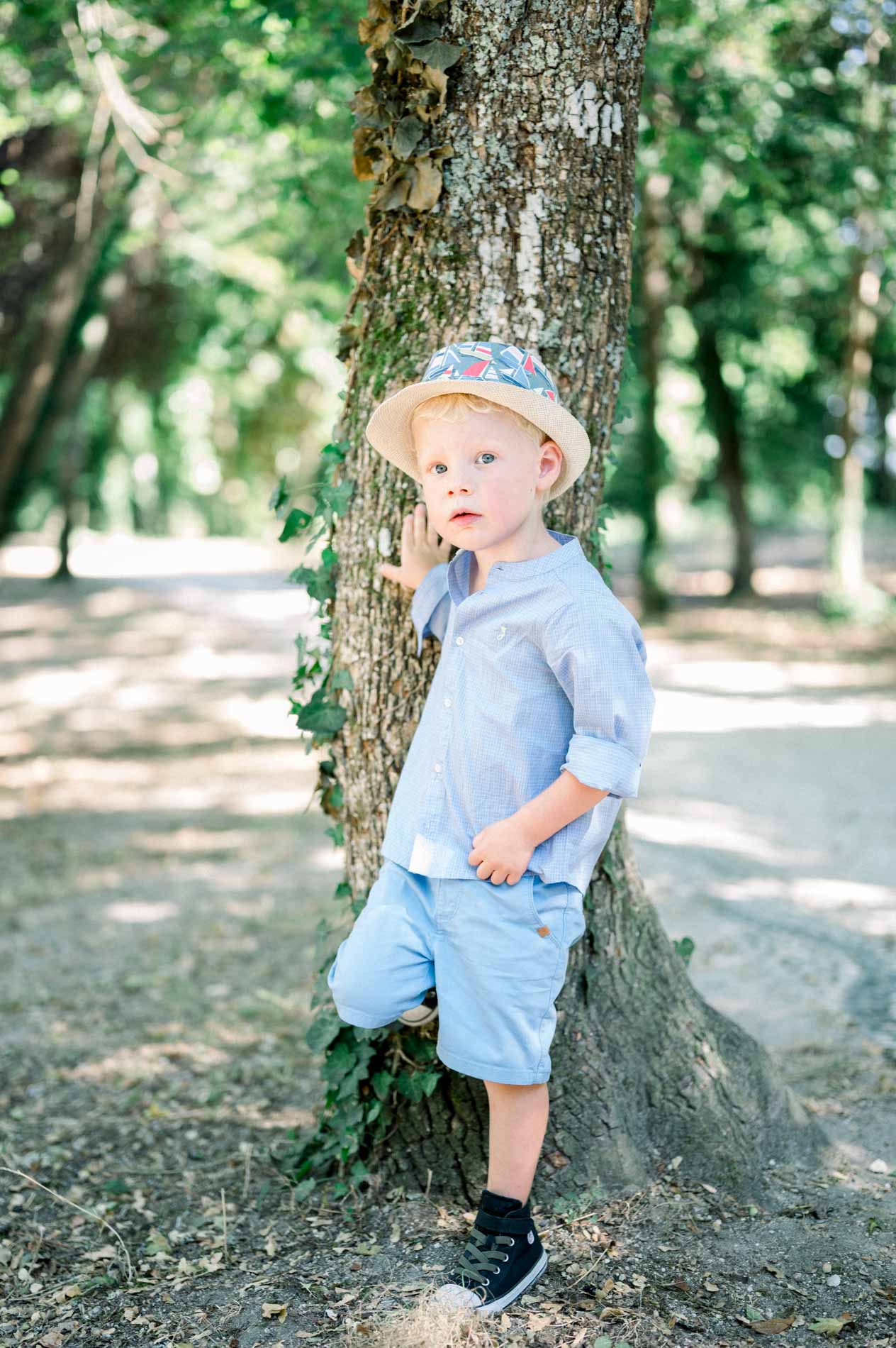 Little blond boy in a blue shirt and hat leaning against a tree trunk