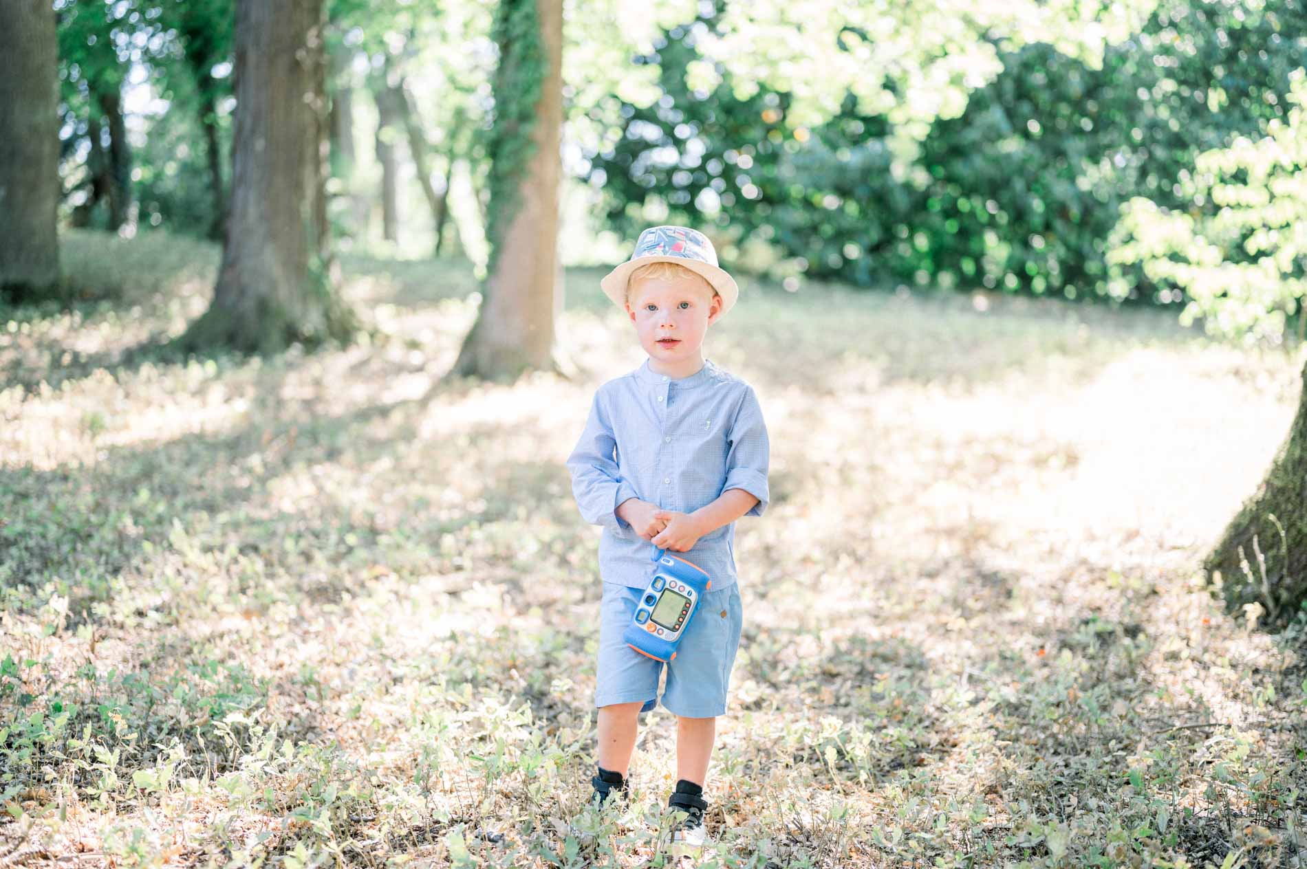 Little boy holding a toy, standing in the grass beneath the trees