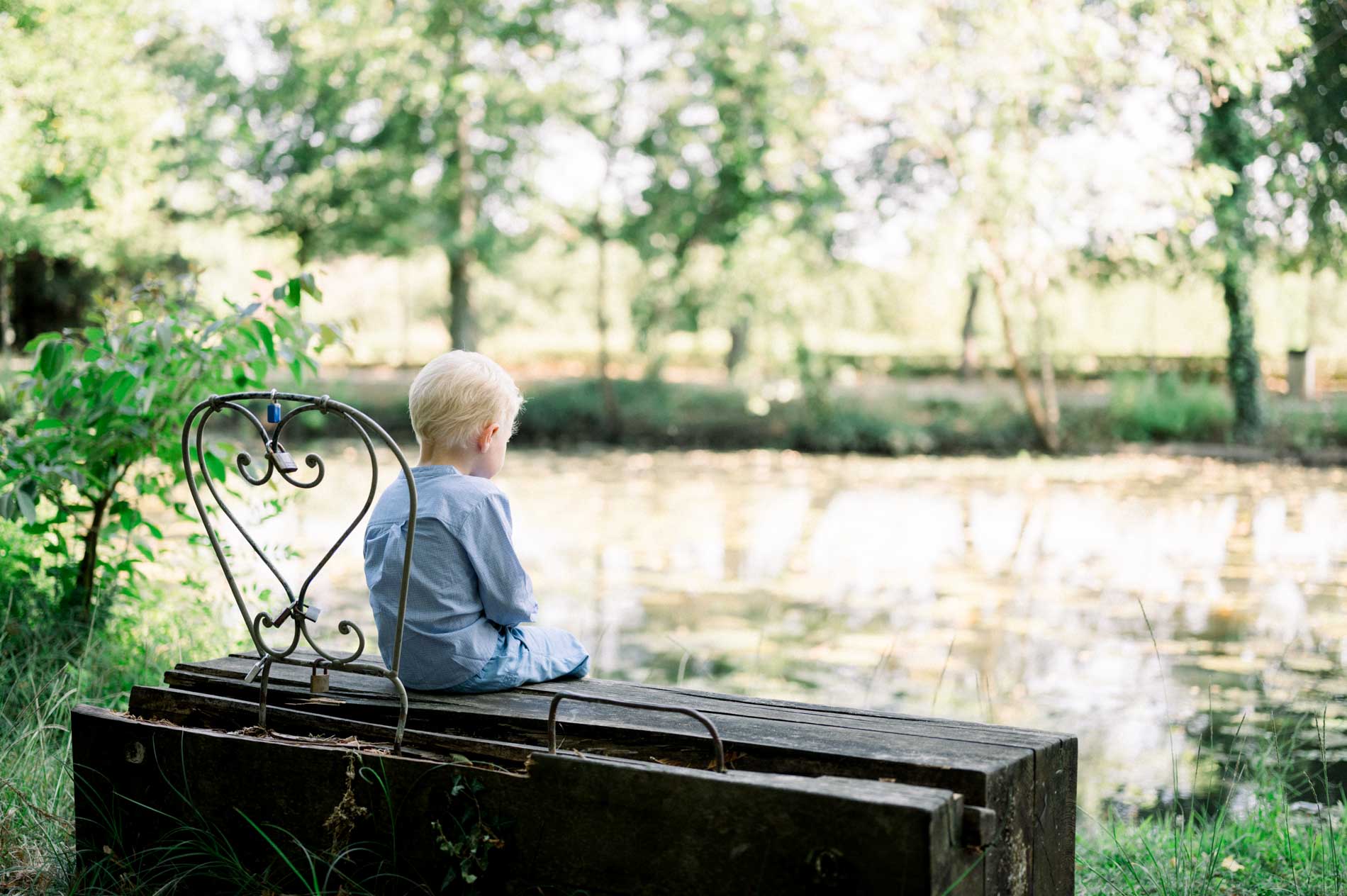 Little boy sitting alone on a bench facing the park lake