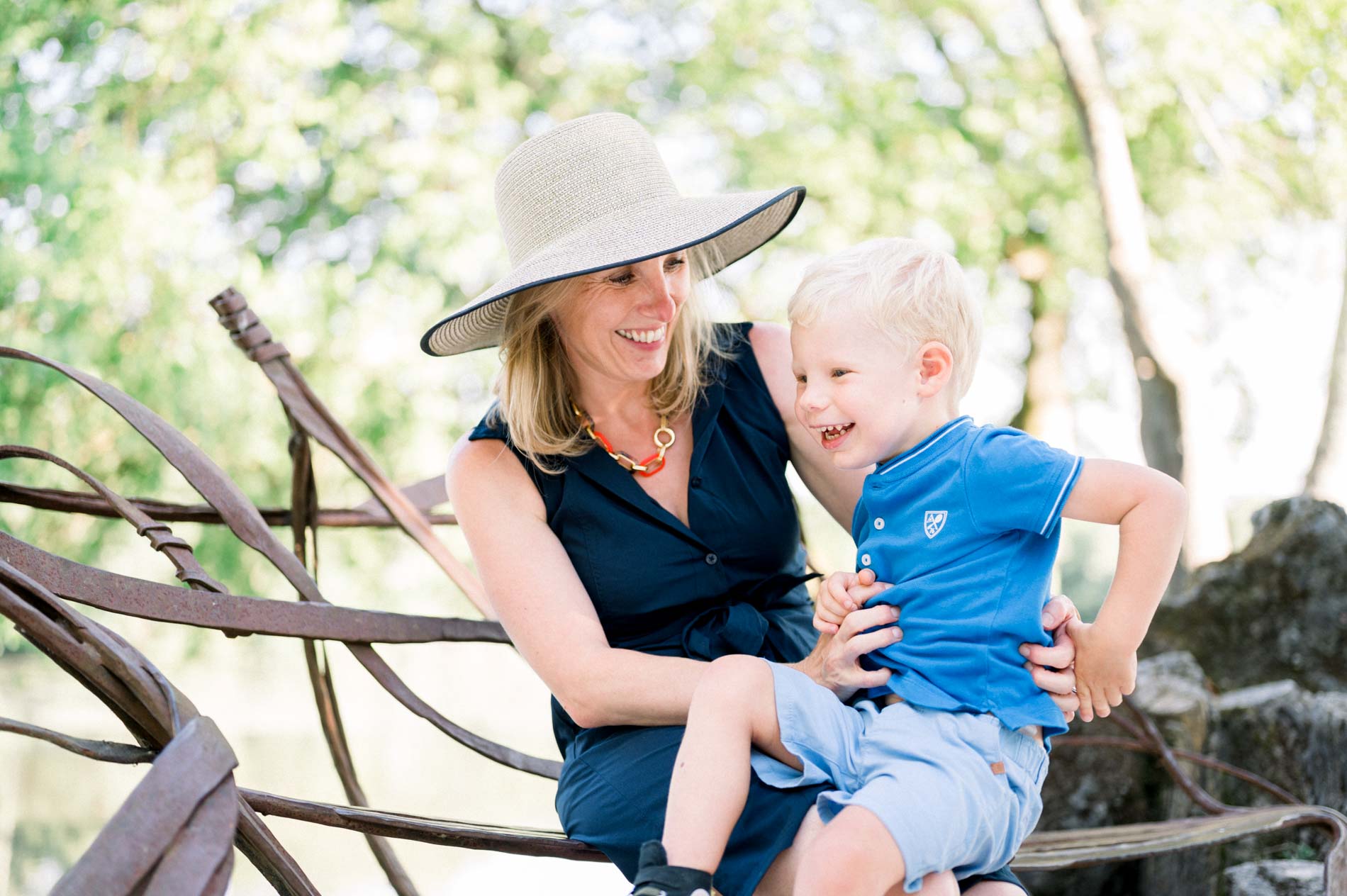 Mum in a straw hat with her little blond son sitting on a wrought-iron rail