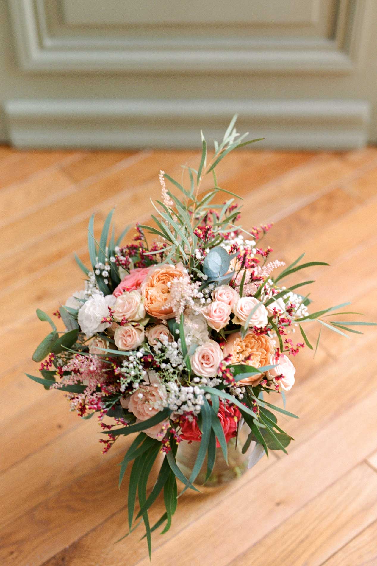 Bouquet de mariée aux roses pêche et feuillage posé sur un parquet clair