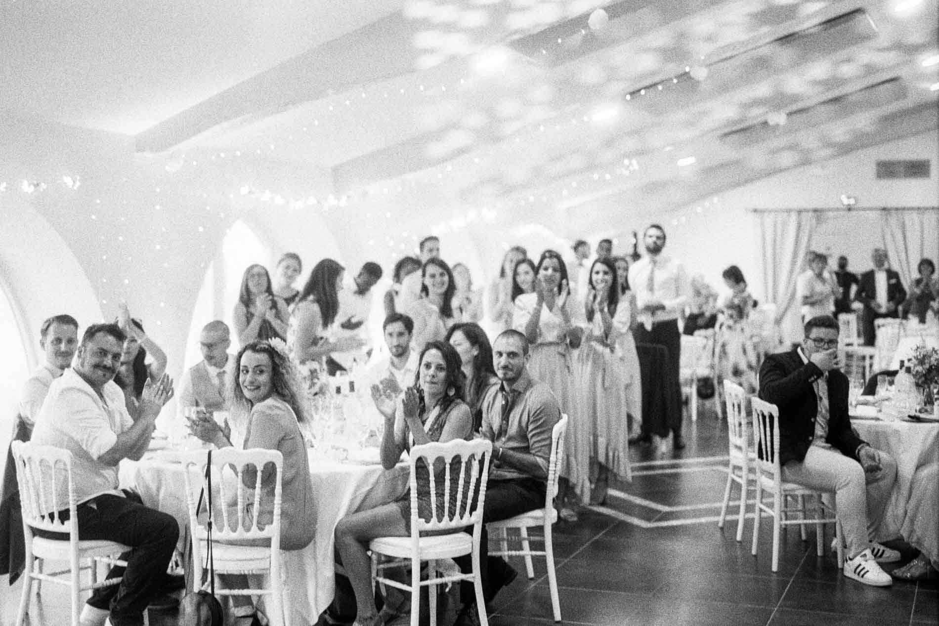 Les invités attablés applaudissent dans la salle de réception, en noir et blanc