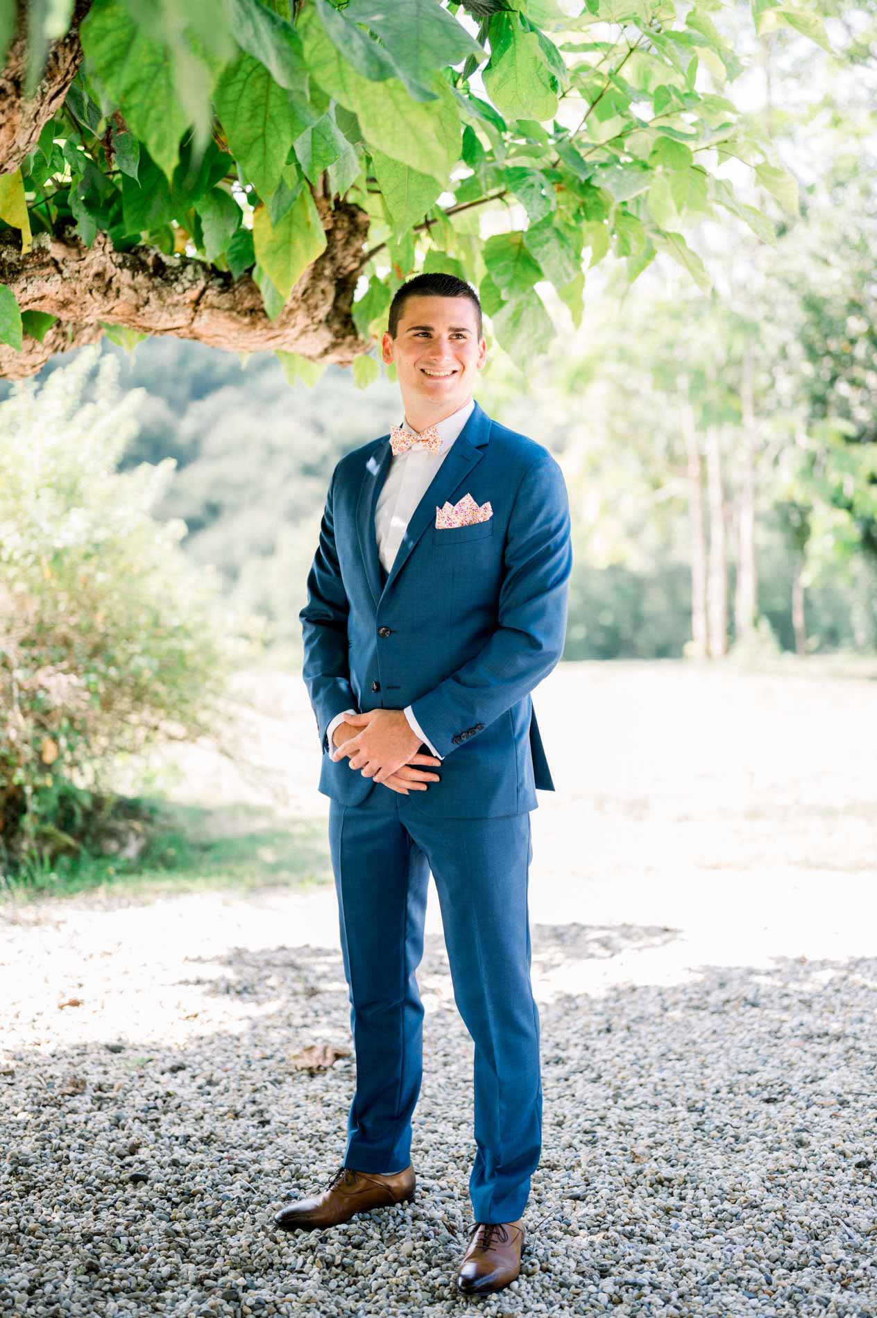 Groom in a blue suit with hands clasped under the trees while getting ready