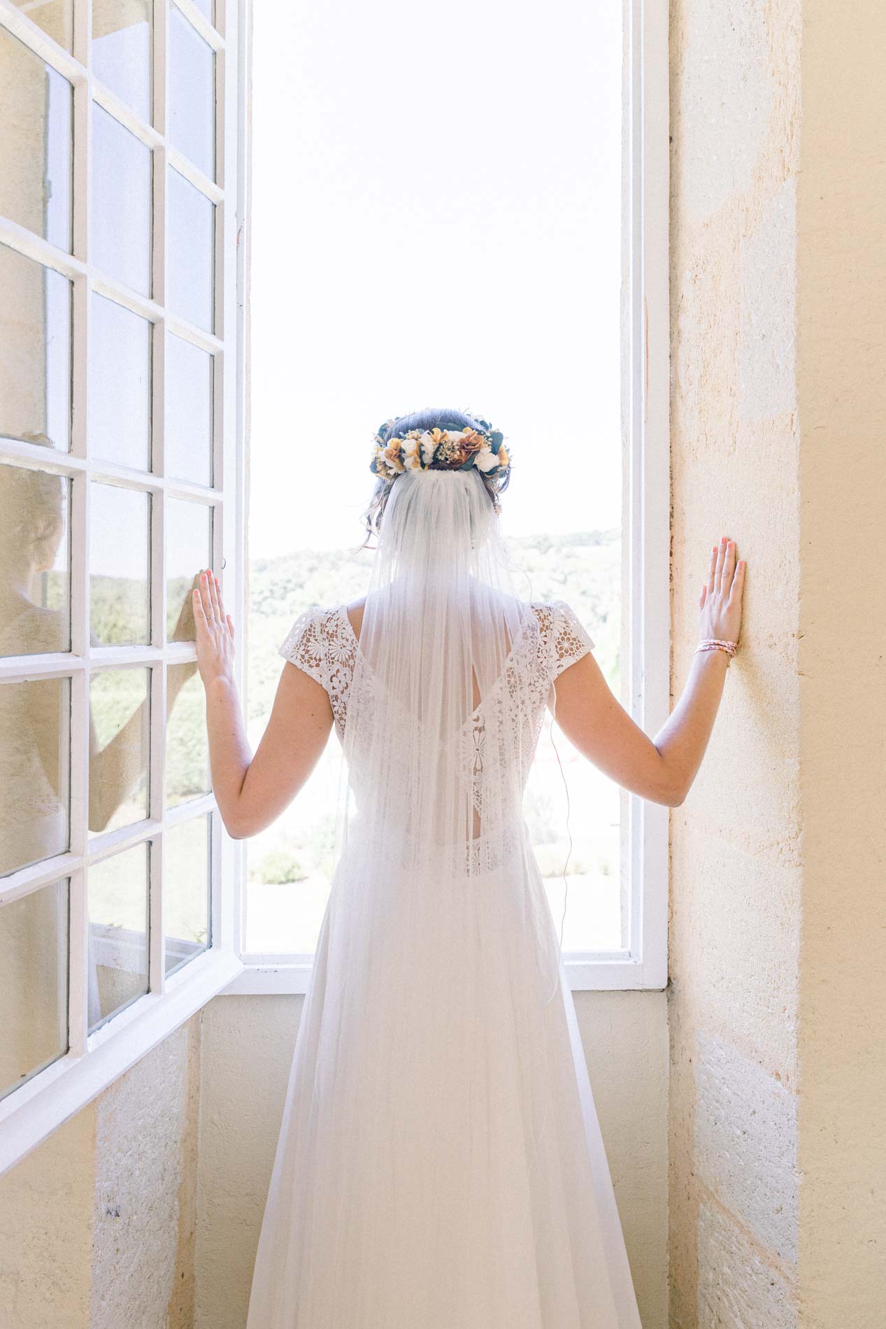 Bride seen from behind, hands resting on the frame of a tall window