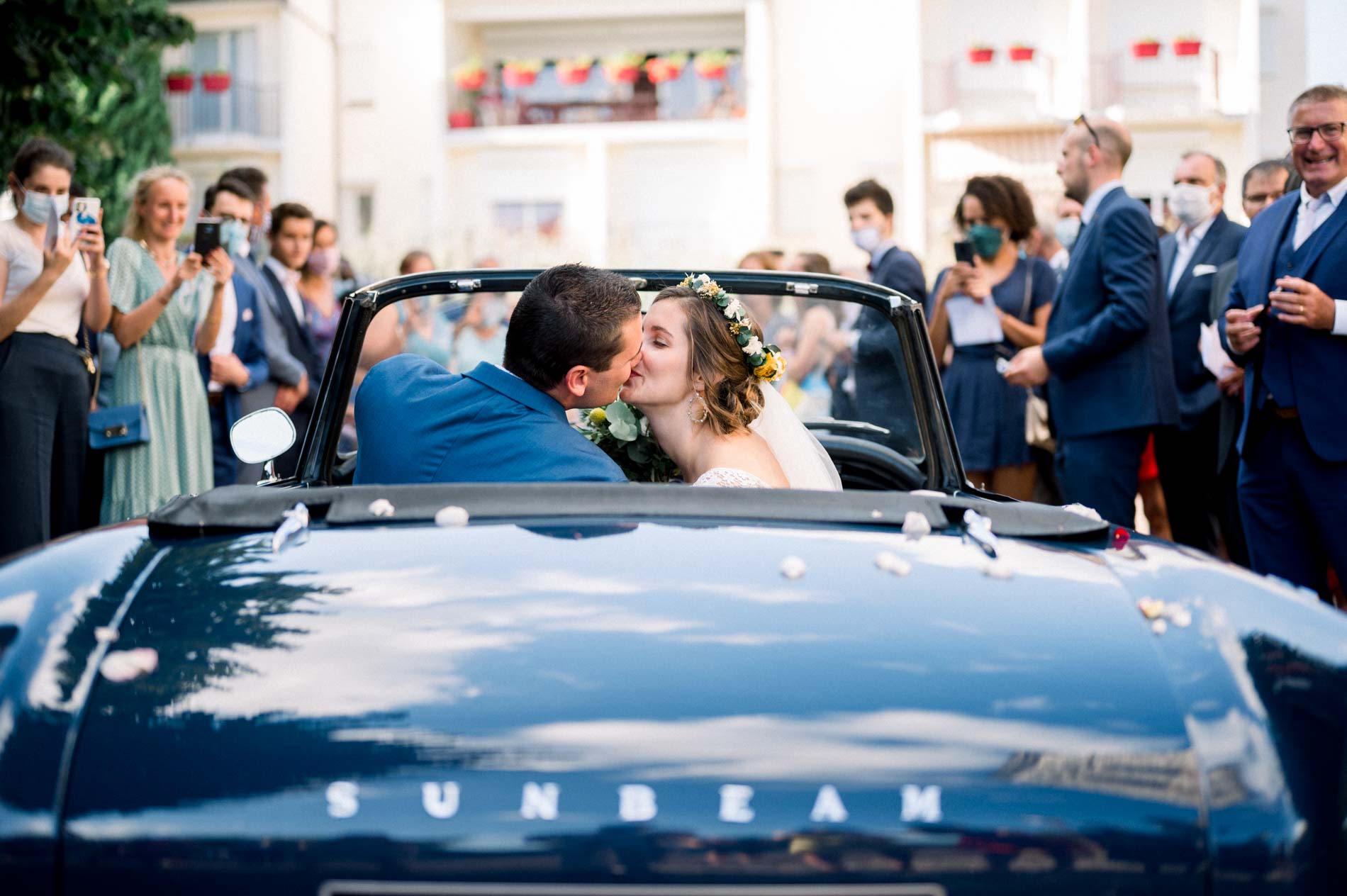 The newlyweds kissing in a blue Sunbeam convertible surrounded by guests