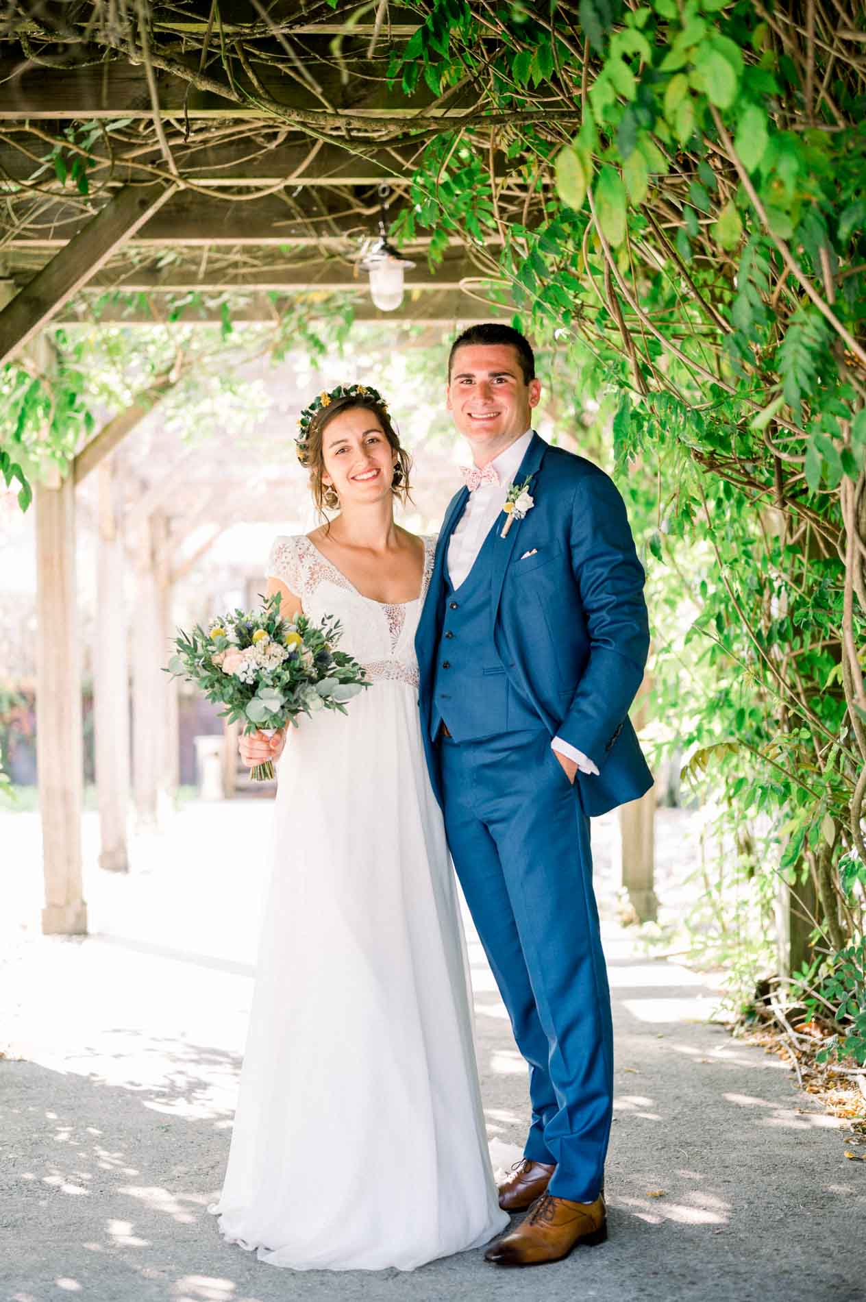 Charlotte and Aymeric under a vine-covered pergola, holding a country-style bouquet