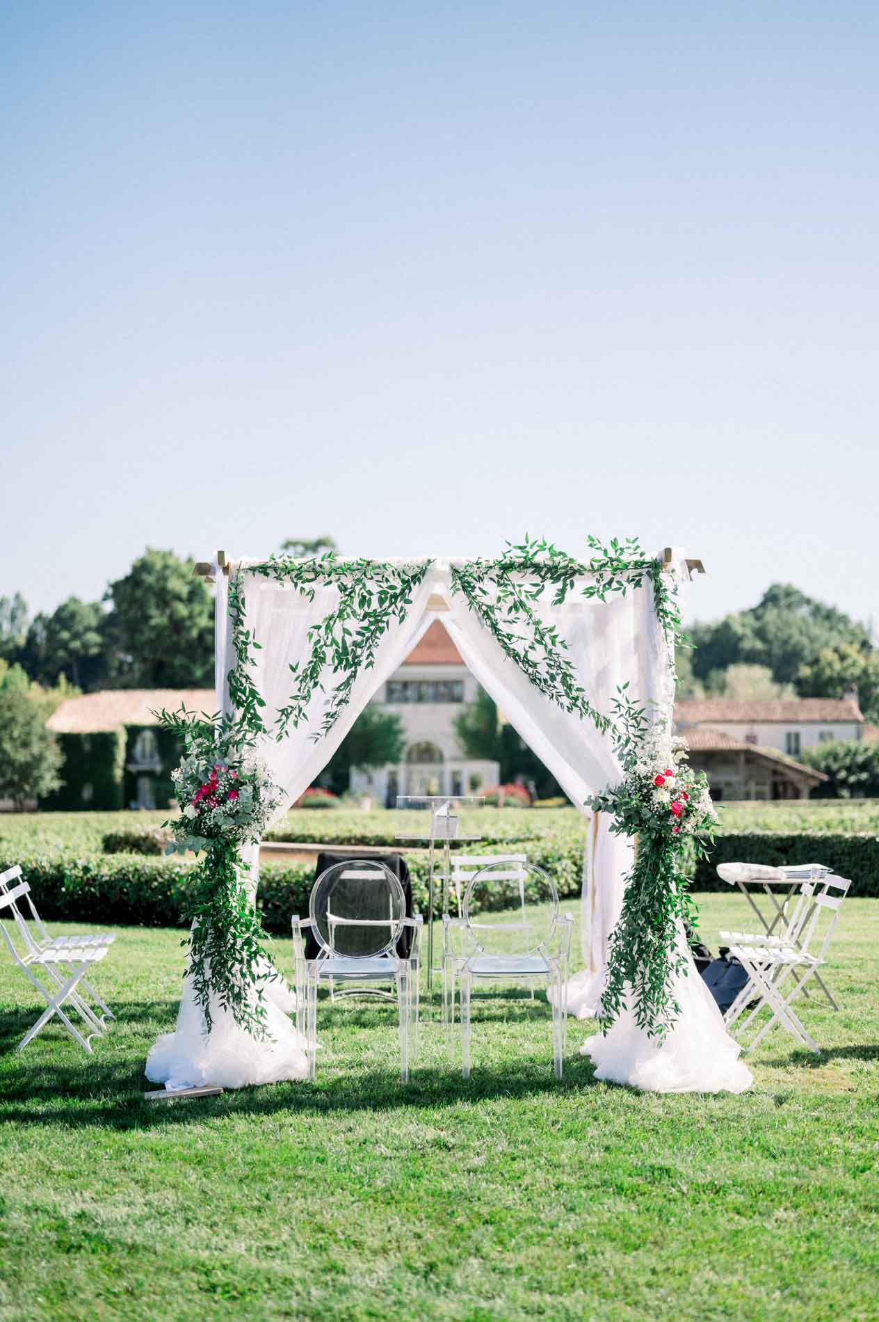 Ecumenical ceremony arch draped with white fabric and greenery on the château lawn