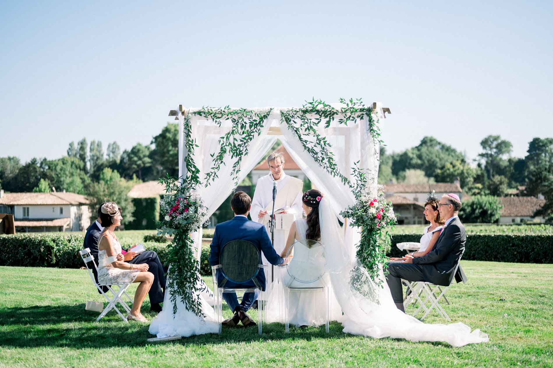 The couple seated beneath the floral arch during the ecumenical ceremony, surrounded by family