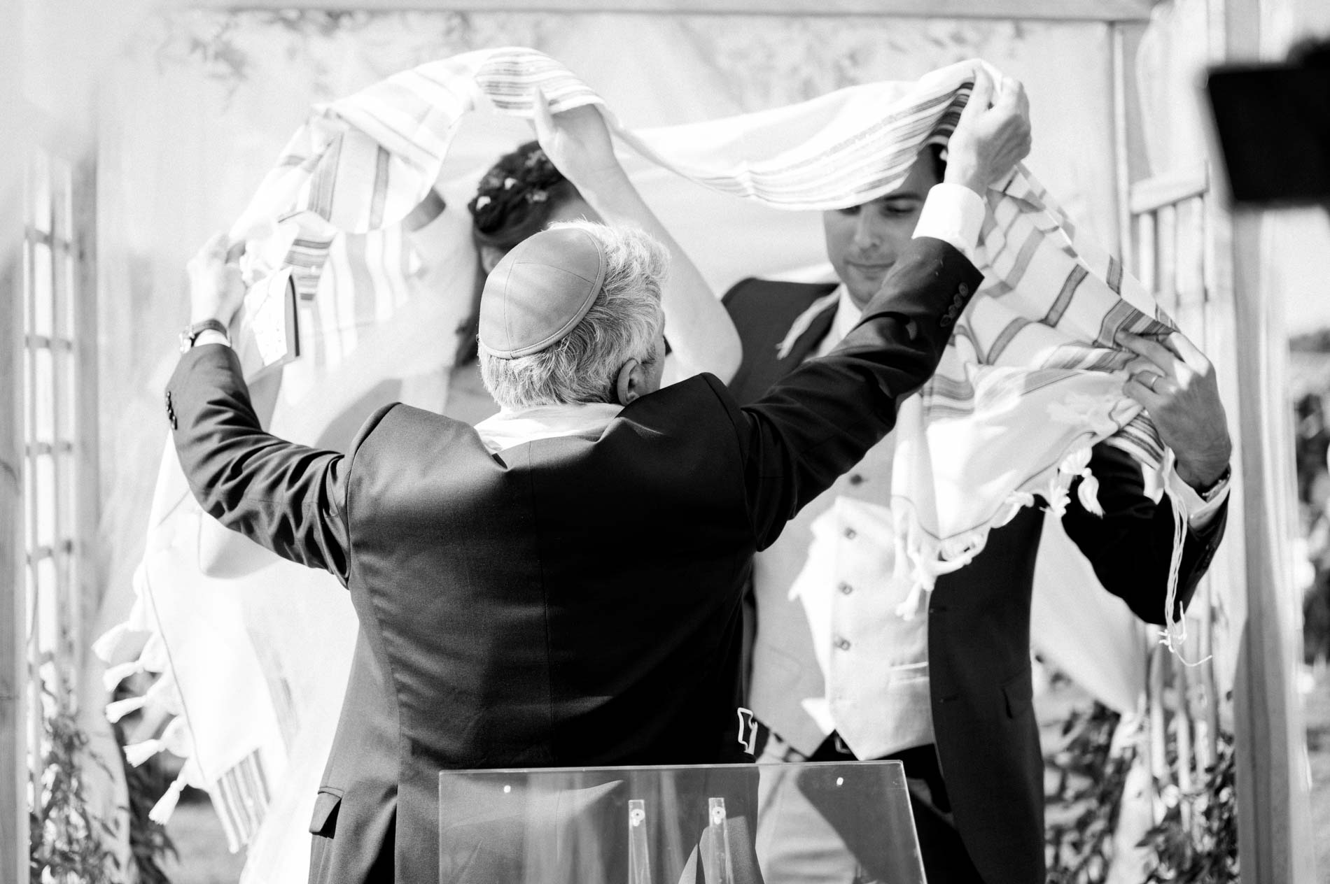 Black and white blessing beneath a prayer shawl held above the couple