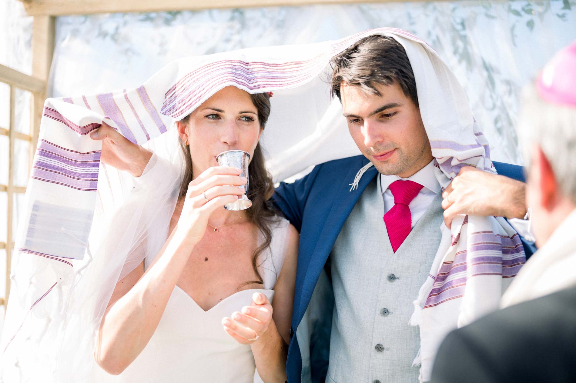 The bride sips wine beneath the prayer shawl beside the groom during the ceremony