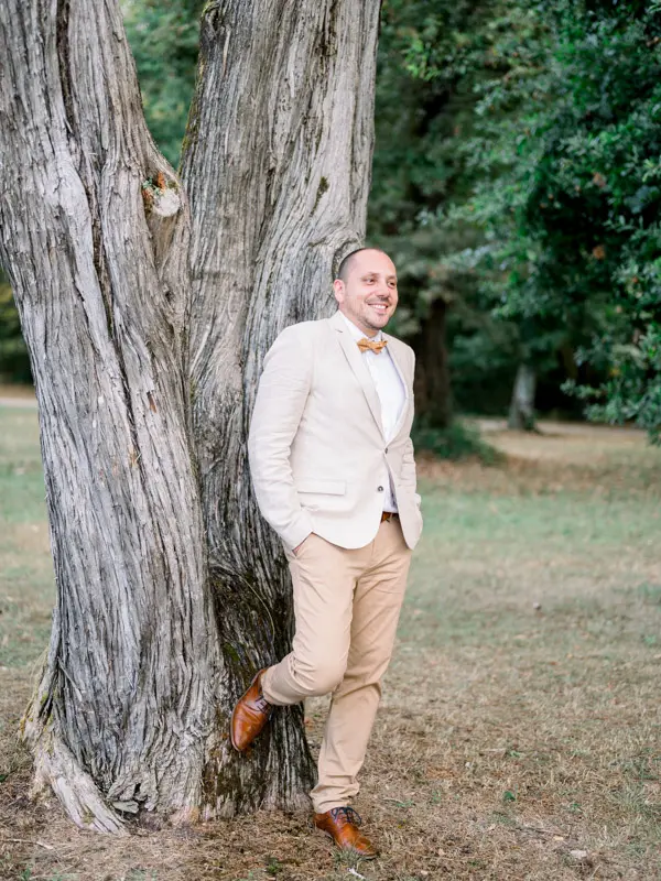 The groom in a linen jacket and bow tie leaning against a large tree in the park
