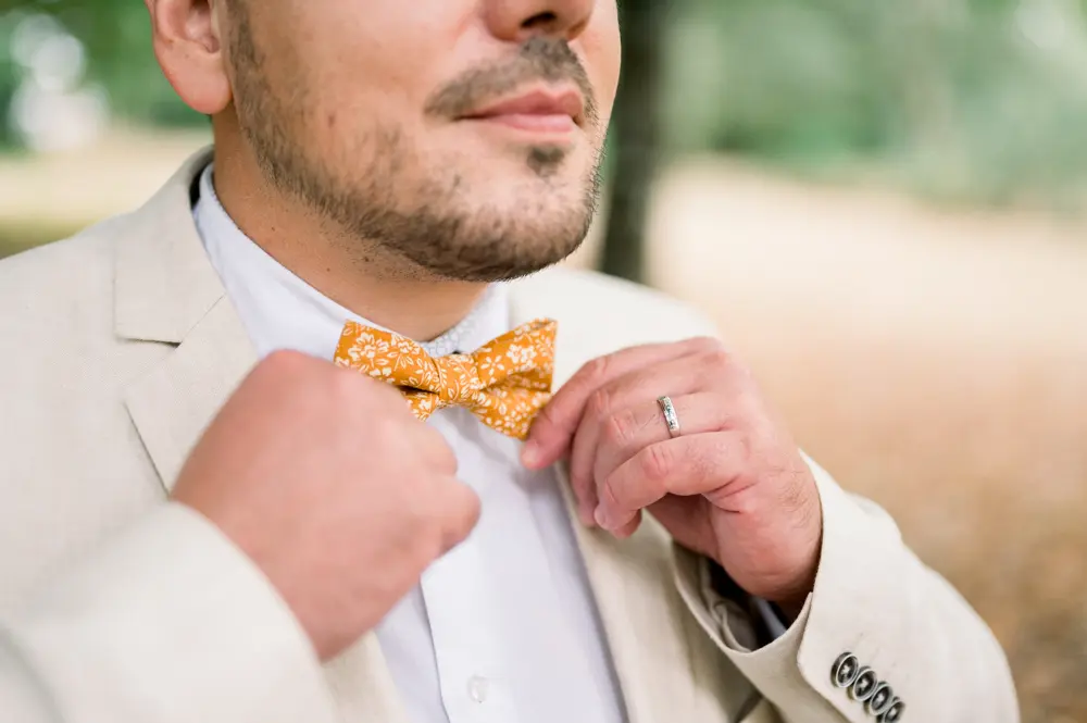 The groom adjusting his mustard bow tie, wedding ring on his finger