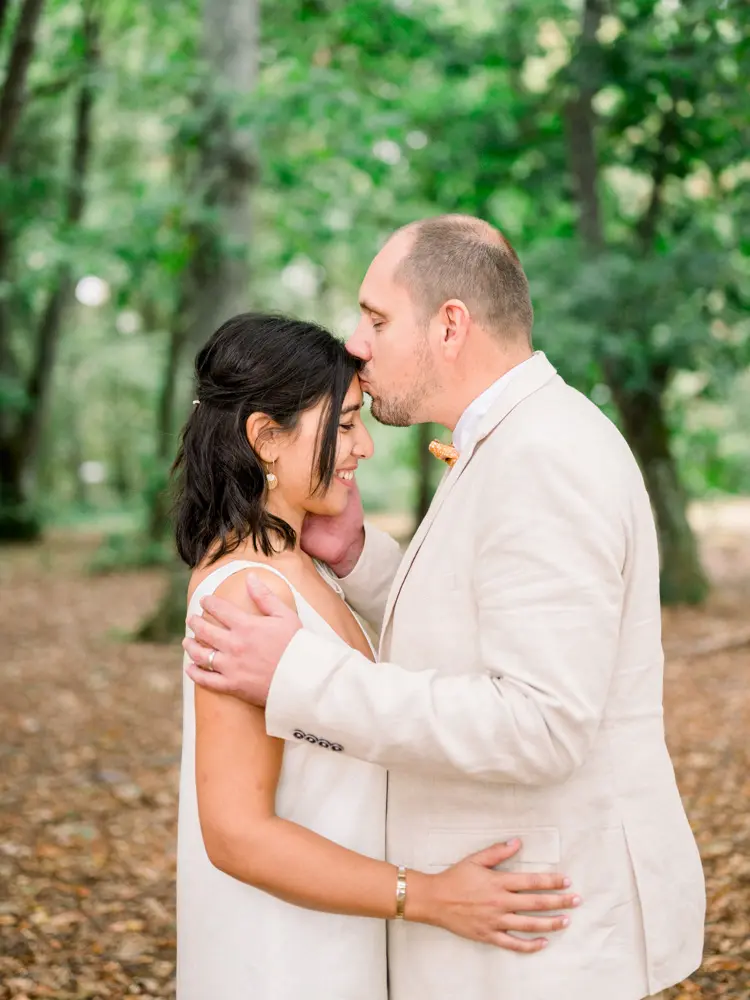 The groom kissing the bride's forehead beneath the trees of the park