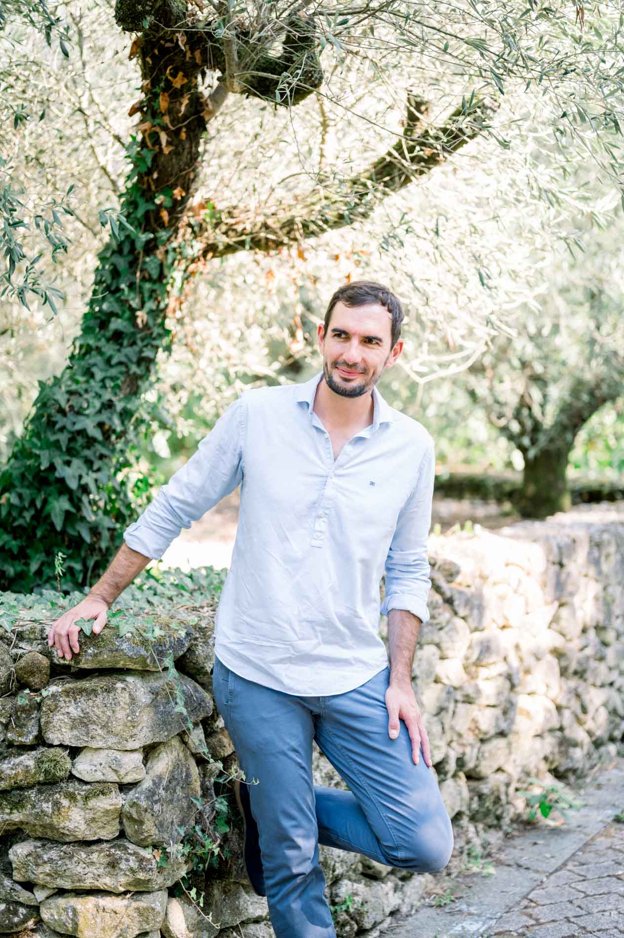 Father-to-be in a blue shirt leaning on a stone wall under the olive trees