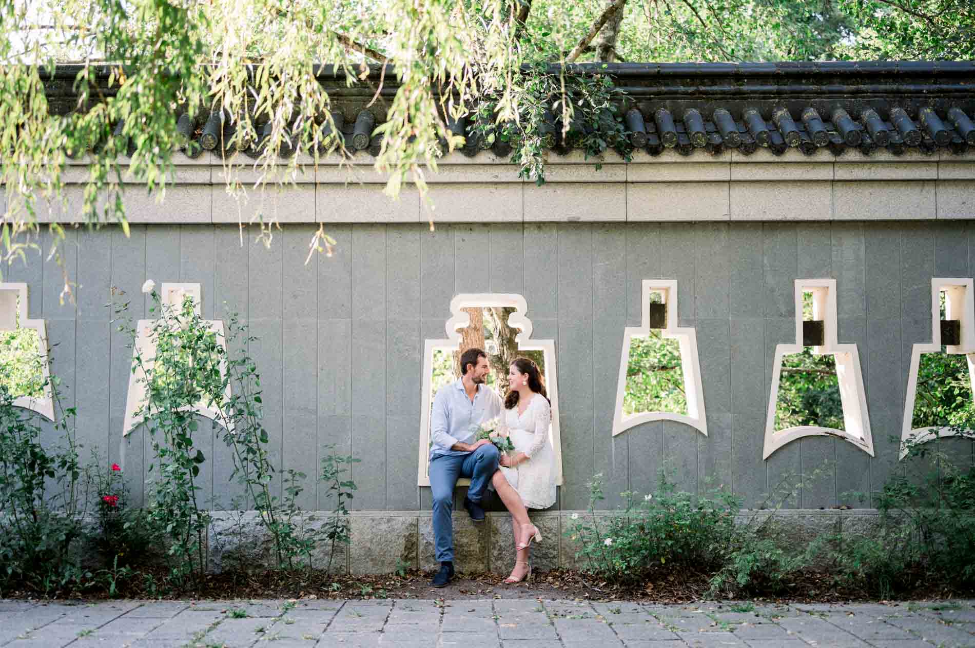Couple sitting before a grey wall with ornamental openings, beneath a willow