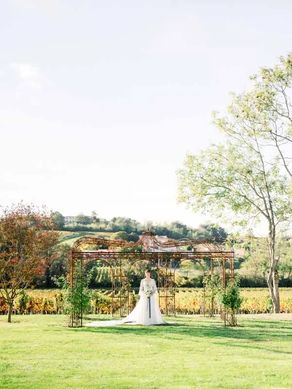 Bride standing alone under a garden arbour facing the autumn vineyards