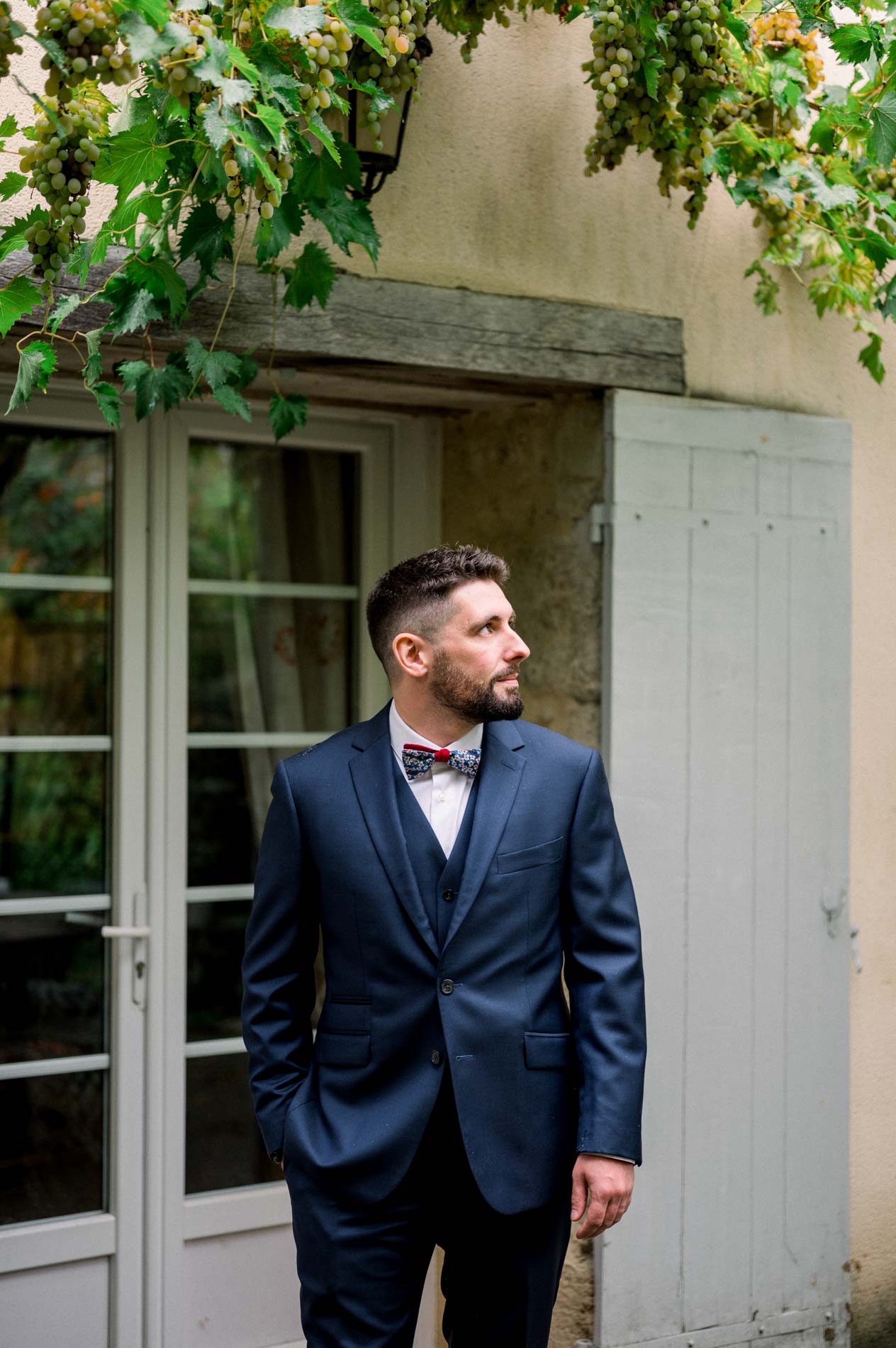 Groom in a navy suit by a French window beneath a vine