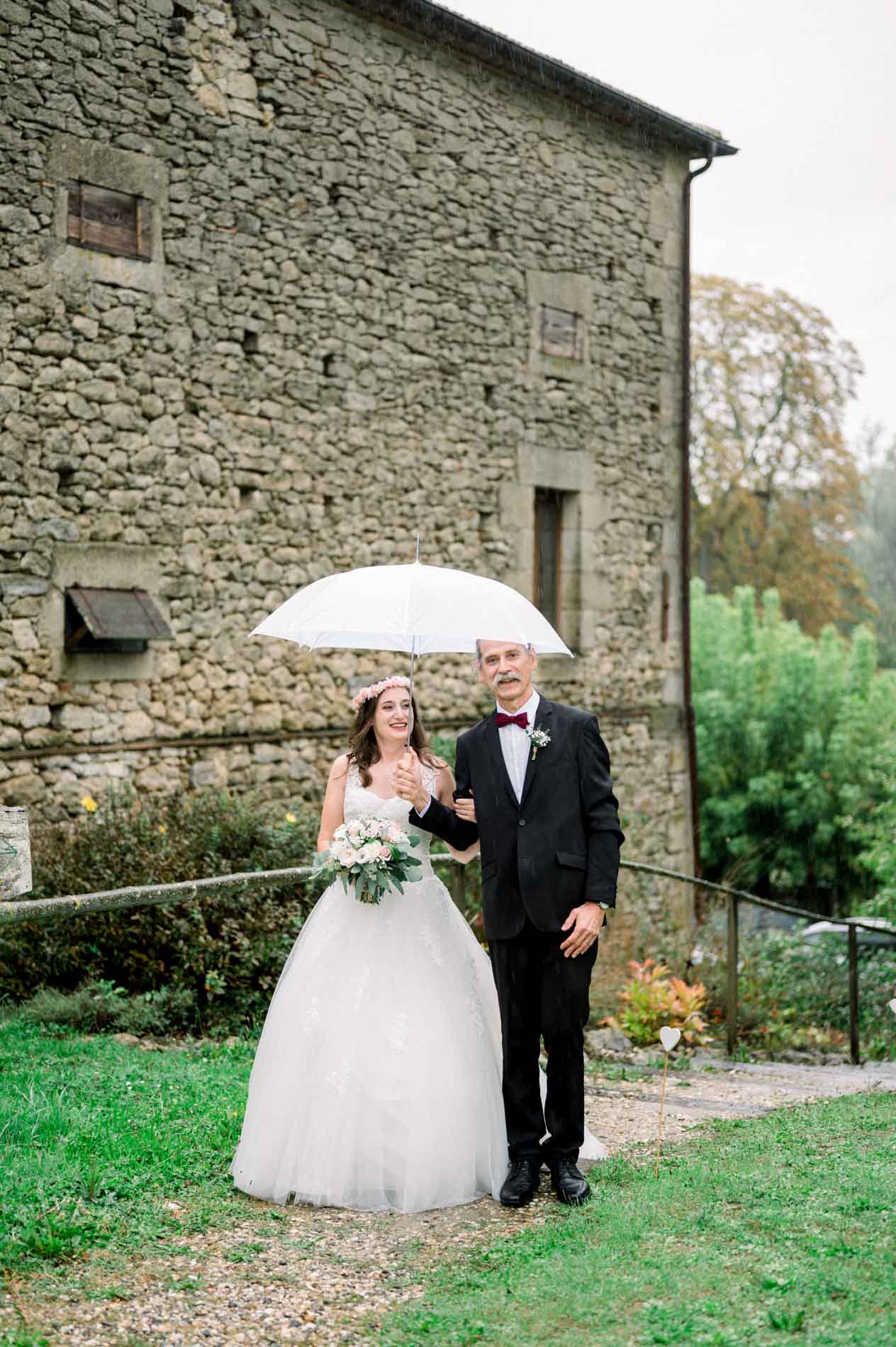 Bride and groom under a white umbrella in front of a stone farm building