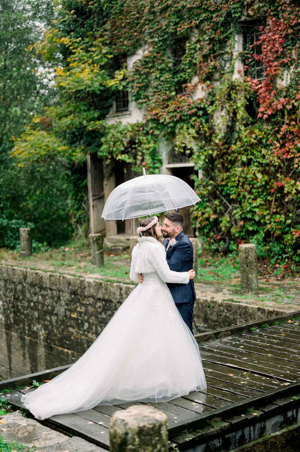 Bride and groom embracing under an umbrella on a wooden footbridge